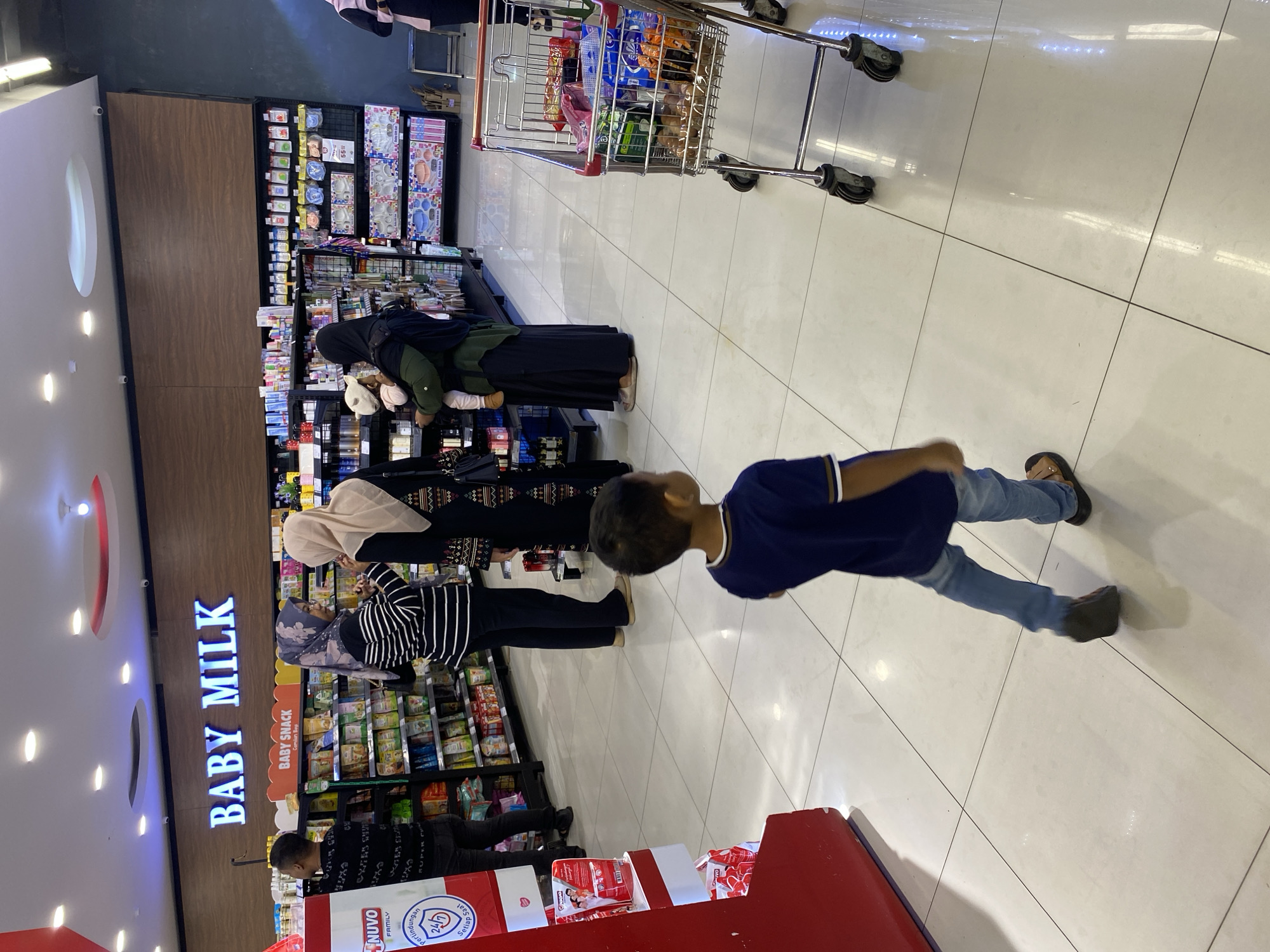 A group of people are shopping in a brightly lit supermarket in Banda Aceh, Indonesia. In the foreground, a young boy in a blue t-shirt and jeans walks away from the camera. Behind him, a shopping cart filled with groceries is parked. Further back, two women, one wearing a black and white striped shirt and the other a beige hijab, are talking. Another woman, wearing a black hijab and green abaya, is holding a baby. A man in a black t-shirt stands near a display of "Baby Milk." The supermarket shelves are stocked with various products, and the floor is tiled and reflective. The overall impression is one of everyday commerce in a modern retail environment.