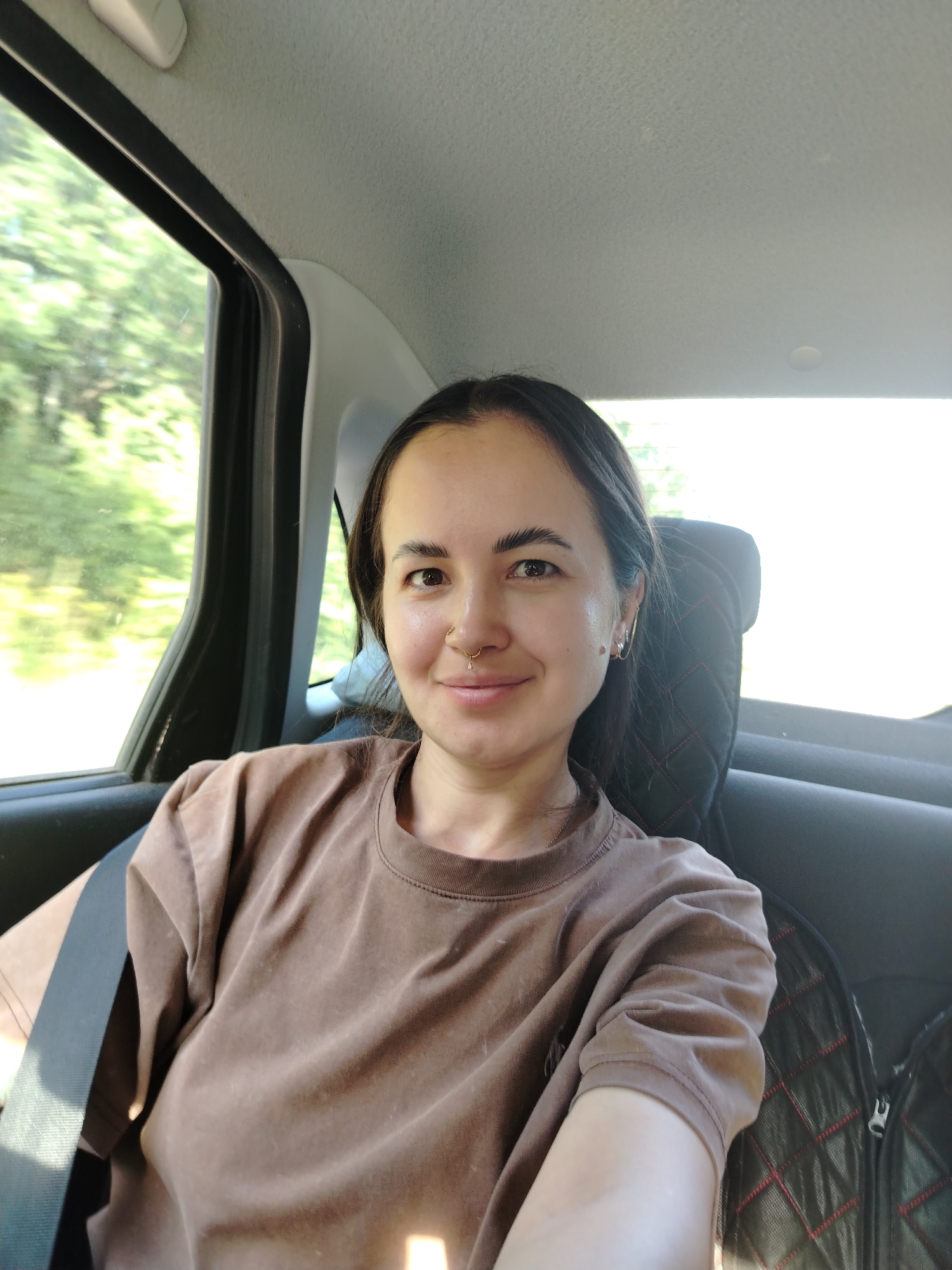 A young woman with dark hair and a nose ring is taking a selfie inside a vehicle. She is wearing a brown t-shirt and a seatbelt. Trees and foliage are visible through the window, suggesting a rural or suburban setting. The interior of the car shows a headrest and part of a seat.