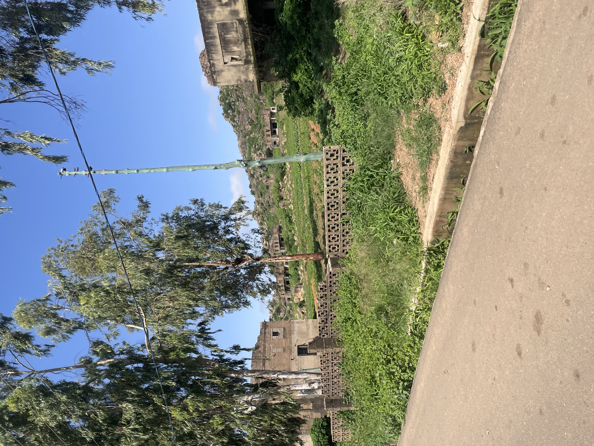 The image depicts an exterior scene in the area of Gura Top, Nigeria, characterized by dilapidated structures and lush vegetation under a clear blue sky. In the foreground, a paved road runs alongside a concrete drainage ditch overflowing with green plants. Behind the ditch, a patterned concrete block wall separates the road from an overgrown area.
Further back, a substantial two-story building with crumbling walls and dark, empty window openings stands partially obscured by tall trees with feathery green leaves. This building appears to be made of stone or concrete blocks. In the mid-ground, the landscape rises into a rocky, green hillside dotted with more ruined structures, some appearing as skeletal frames of buildings. A tall, slender green pole, possibly a utility pole, stands vertically in the middle ground, supporting a wire that stretches across the frame. The overall impression is one of abandonment and nature reclaiming man-made structures in a semi-arid environment.