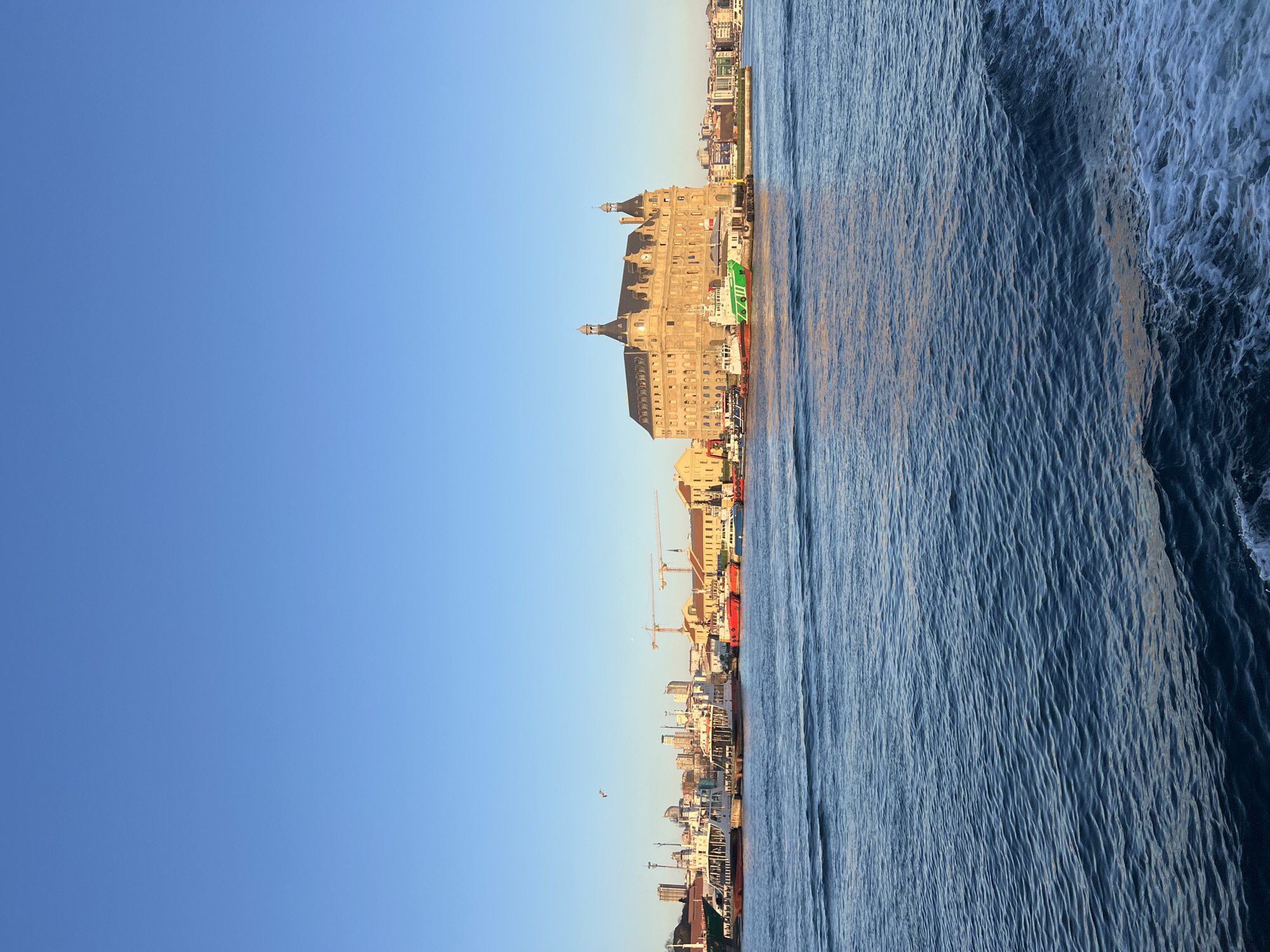 The image presents a view of the Haydarpaşa Terminal, a railway terminal located in Kadıköy, Istanbul, Turkey, viewed from the sea. The terminal is a large, ornate building with a distinctive architectural style. Adjacent to the terminal is a harbor with several ships docked. Behind the terminal and harbor lies a section of the city, revealing buildings, cranes, and other urban infrastructure. The sea is rough with visible waves. The sky is clear with a gradient of blue.