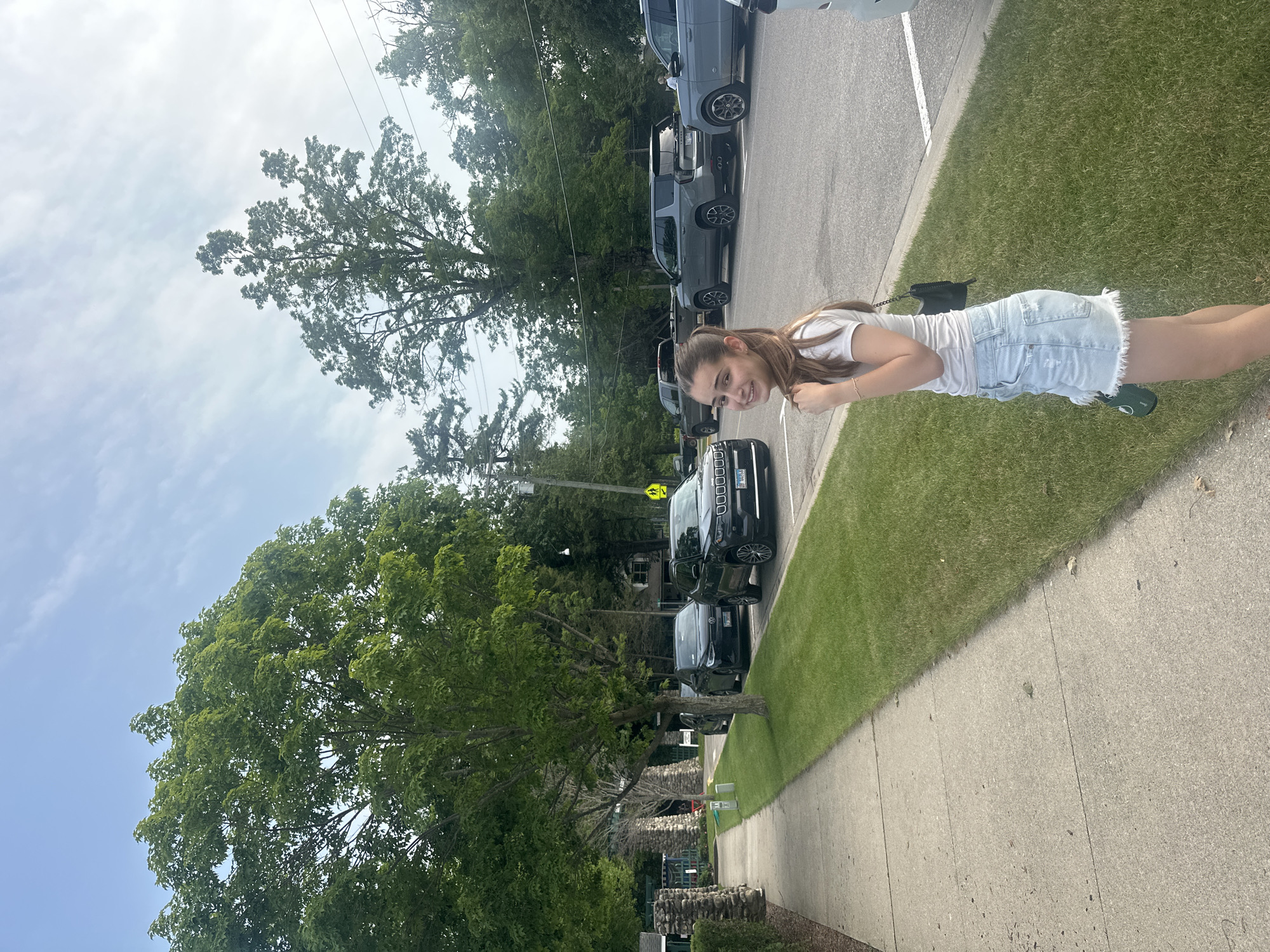 A young woman with long brown hair, wearing a white t-shirt and light blue denim shorts, stands on the grass next to a sidewalk in Harbor Springs, Michigan. She is smiling at the camera and holding her hair. Behind her, cars are parked along a street, and mature trees with green foliage line the sidewalk and parking area. The sky is partly cloudy with a soft, natural light. In the background, there appears to be a stone structure and some buildings, suggesting a park or a well-maintained street.