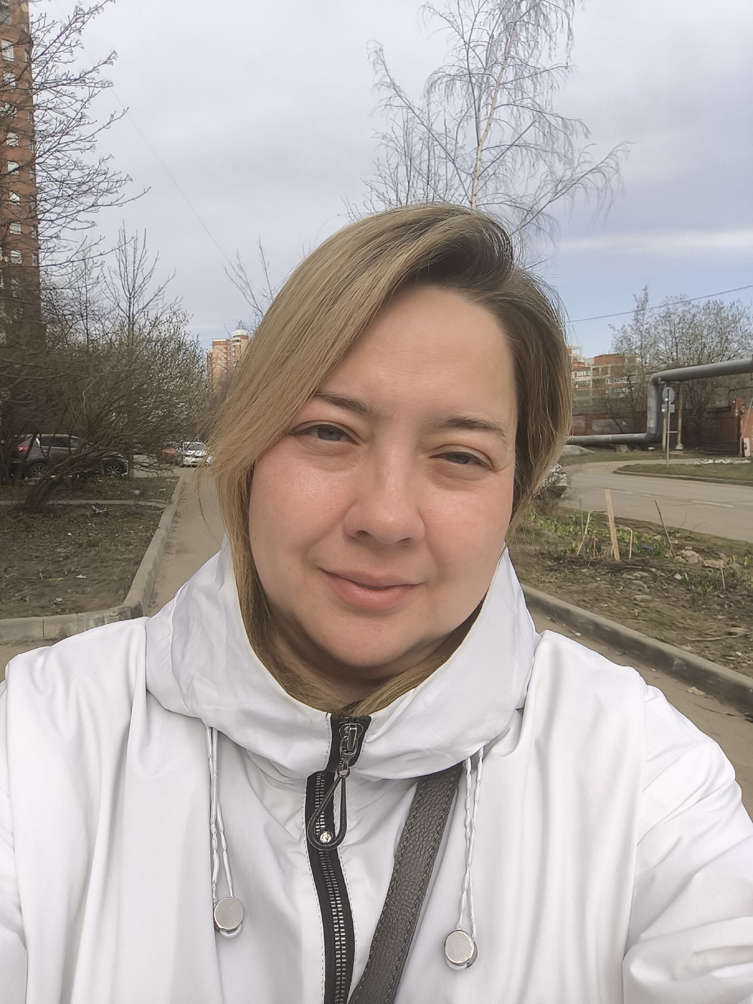 A woman with blonde hair, wearing a white hooded jacket, is taking a selfie outdoors in Khimki, Russia. She is standing on a sidewalk next to a road. In the background, there are buildings, trees with bare branches, and a complex of pipes. Several parked cars can be seen. The sky is overcast.