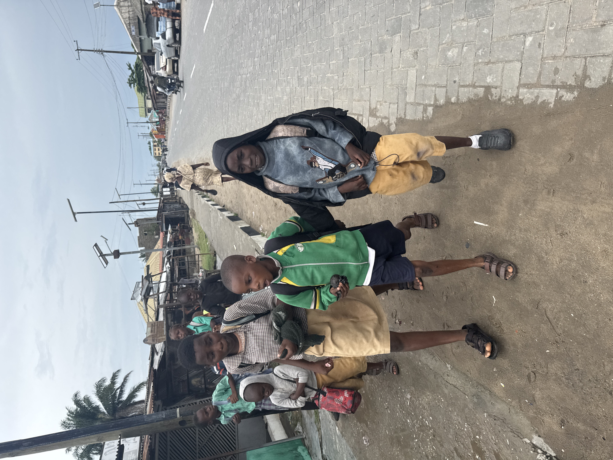A group of African children are walking and standing on a paved road in Badagry, Nigeria. The road is lined with buildings and a few cars in the background. The sky is overcast. The children appear to be of school age and are wearing various casual clothes. Some of them are holding objects in their hands.