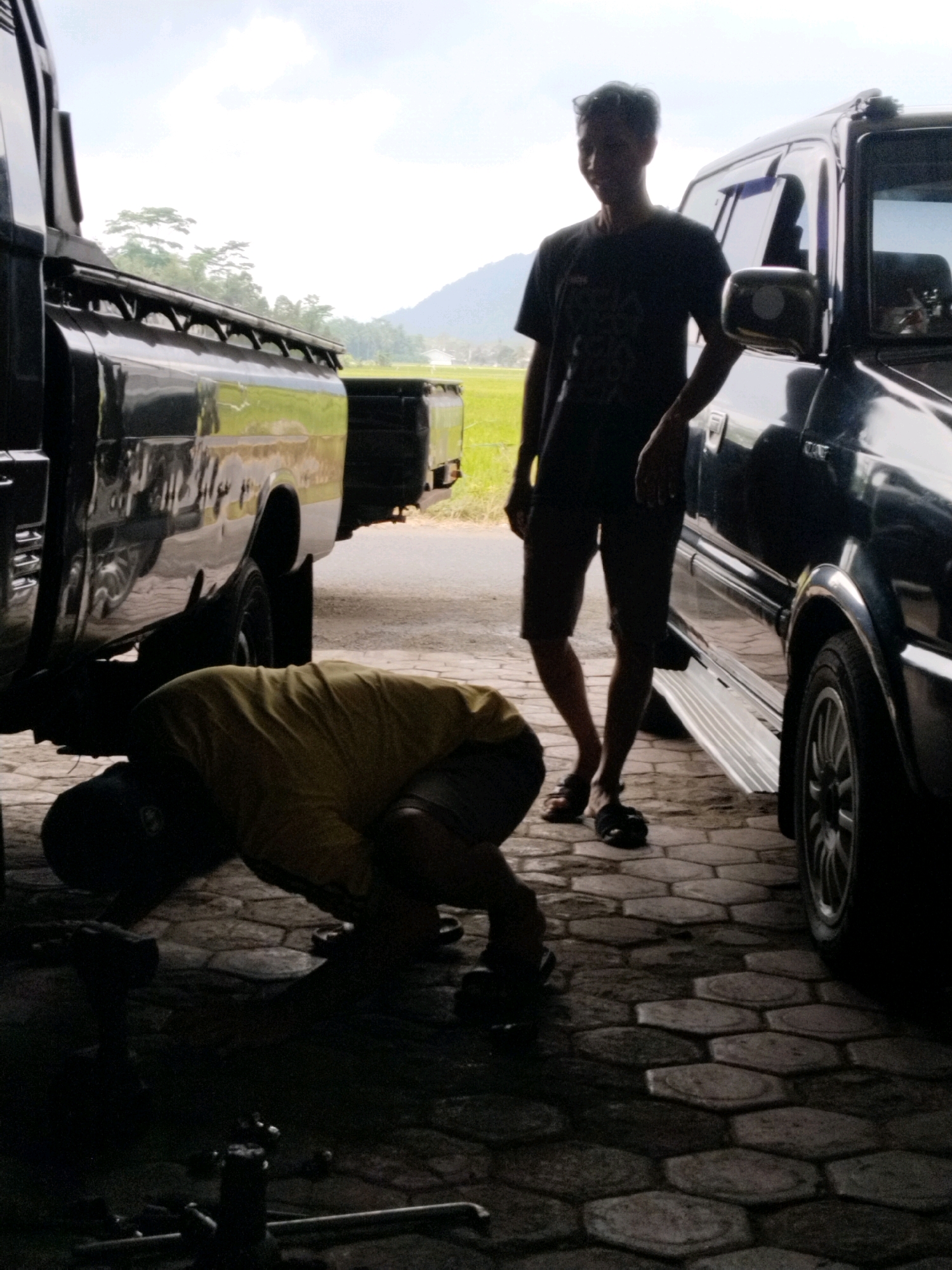 The image captures a dimly lit outdoor scene in Prigi, Indonesia, likely a repair shop or garage. Two individuals are present. One, silhouetted and wearing a dark t-shirt and shorts, stands near a black SUV, looking towards the camera. The other individual, also silhouetted and wearing a yellow t-shirt and dark shorts, is crouched down, seemingly working underneath a black pickup truck. The ground is paved with hexagonal stone tiles. In the background, a lush green field and distant hills are visible under a bright, cloudy sky, suggesting an open, rural setting. The lighting conditions create strong shadows, obscuring finer details of the faces and immediate surroundings of the subjects.