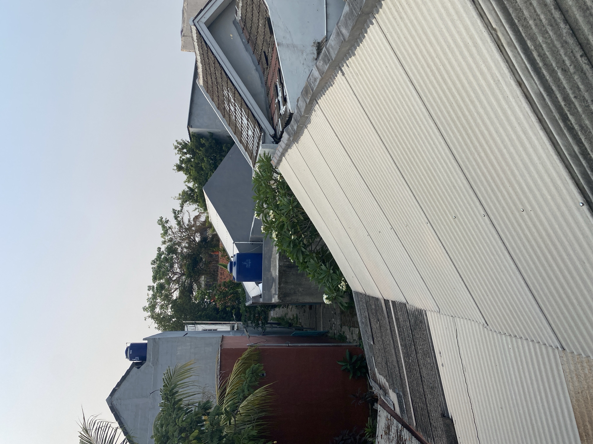 The image depicts a view of rooftops and buildings in a residential area of Tangerang, Indonesia. In the foreground, a corrugated metal roof occupies the lower right portion of the frame, transitioning to an older, darker corrugated roof on the left. Behind these roofs, several buildings of varying heights and architectural styles are visible.
To the left, a tall, narrow building with a red facade and a grey upper section stands prominently. A blue water tank is mounted on the roof of a grey building to the right of this red building. In the mid-ground, a narrow pathway or alleyway winds between structures. Lush green trees and foliage are interspersed throughout the scene, indicating a tropical or subtropical environment. The sky above is clear and pale, suggesting daytime, likely early morning or late afternoon due to the soft lighting. The overall impression is of a densely populated urban or suburban area characterized by a mix of modern and older construction.
