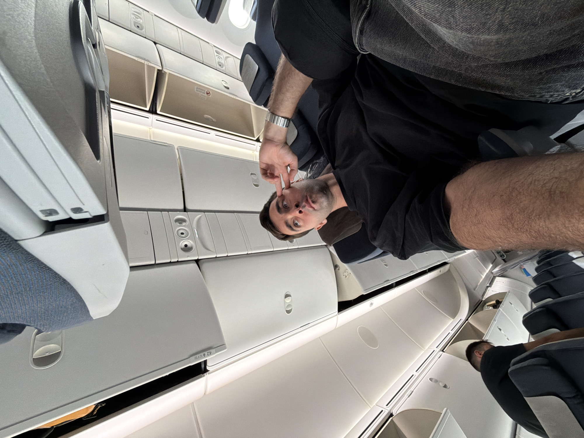 The image shows the interior of an airplane cabin. A man is seated, facing the camera. He is wearing a black t-shirt, gray jeans, and a silver watch, and he is making a face with his fingers on his cheeks. Overhead compartments are visible above the seats. In the background, other seated passengers can be seen. The image was likely taken onboard a flight departing from or arriving at Phuket International Airport, which is located in Tambon Mai Khao, Thailand.