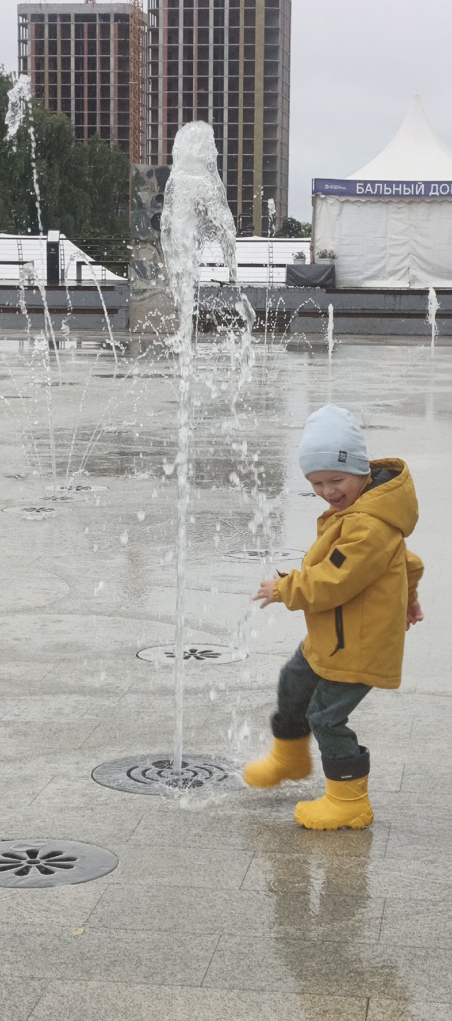 This image depicts a young boy playing in a public fountain in Izhevsk, Russia. The boy, wearing a yellow hooded jacket, jeans, and yellow rubber boots, is mid-stride, with his mouth open in a laugh and his arms outstretched. He is positioned in the foreground, with a tall water spout from the fountain directly in front of him, creating a spray that partially obscures him and the surrounding area.
The fountain is set in a paved plaza with a stone-like texture. Circular grates are visible in the pavement, indicating the water jets. The ground is wet and reflective, showing the water's presence. In the background, several buildings are visible. To the left, two tall, unfinished buildings with visible rebar and scaffolding suggest ongoing construction. To the right, a large white tent with a blue banner that reads "БАЛЬНЫЙ ДОМ" (Ballroom) is set up, hinting at an event or public gathering space. Trees are also visible behind the buildings, adding a touch of greenery to the urban landscape. The sky is overcast, contributing to the subdued lighting of the scene.