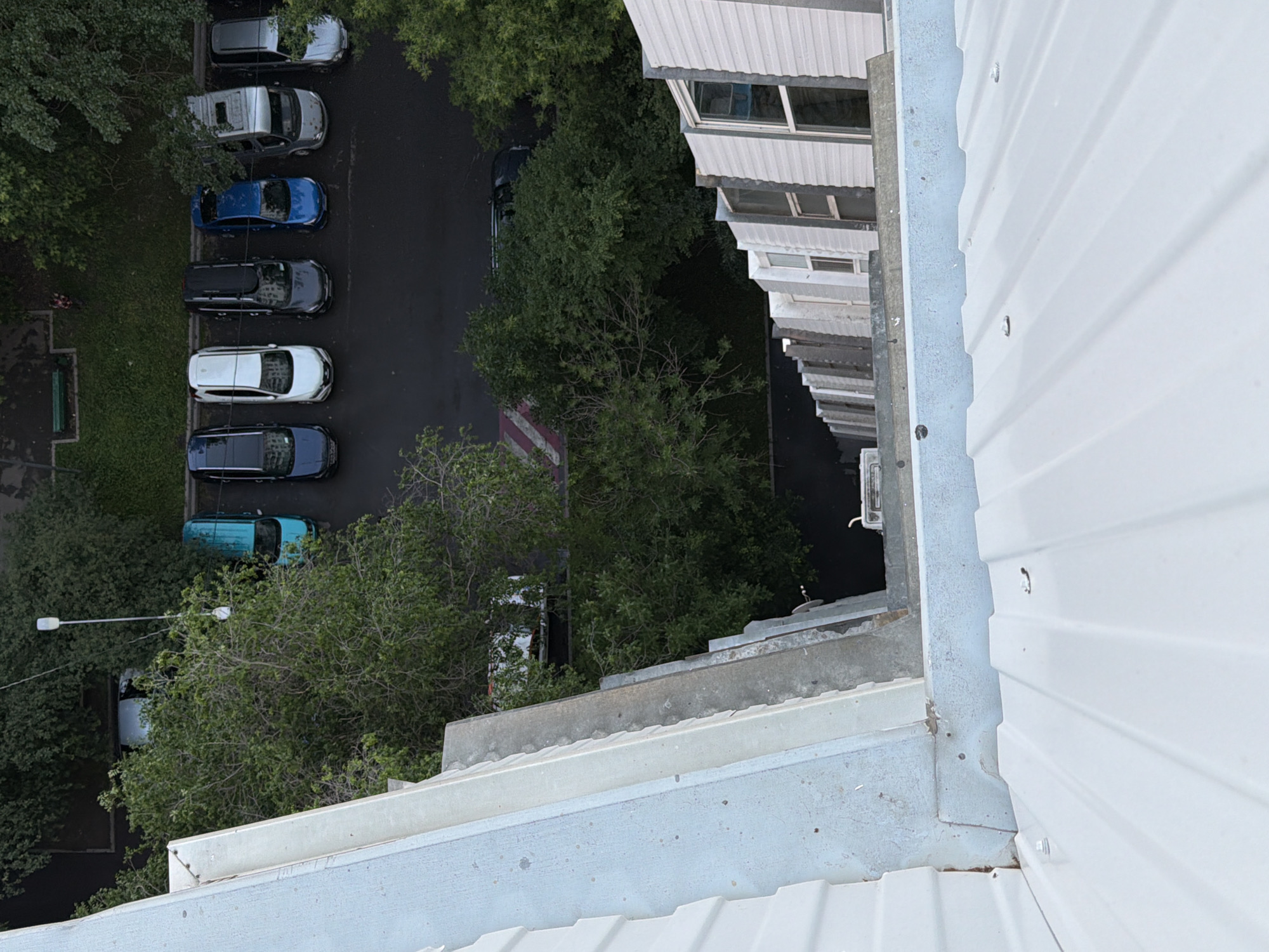 The image, taken from a high vantage point, shows a residential area in Khimki, Russia, with a row of parked cars visible on a street. The cars, including a blue hatchback, a white sedan, two dark-colored SUVs, a blue minivan, and a silver minivan, are parked in a line. Trees and buildings with multiple windows are also present in the background. The foreground of the image is dominated by the roof of a structure, which appears to be made of white corrugated metal. The scene suggests a typical urban setting with residential buildings and street-level activity.