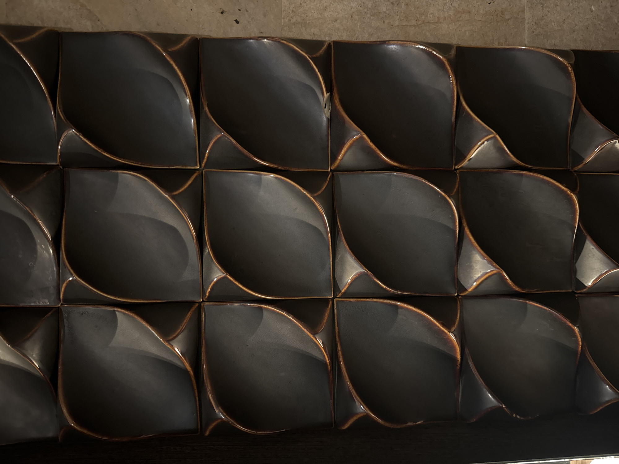 A close-up shot presents a wall composed of dark, possibly black or very dark gray, tiles with a distinctive three-dimensional, curved design. The tiles are square and arranged in a grid pattern. The edges of the tiles have a visible brown glaze or finish, creating a defined border around the darker centers. The lighting in the scene is uneven, casting shadows that accentuate the curved surfaces of the tiles and create a contrast between light and dark areas. The overall impression is of a modern or decorative wall covering. The scene is located in the city of Pecatu, Indonesia.