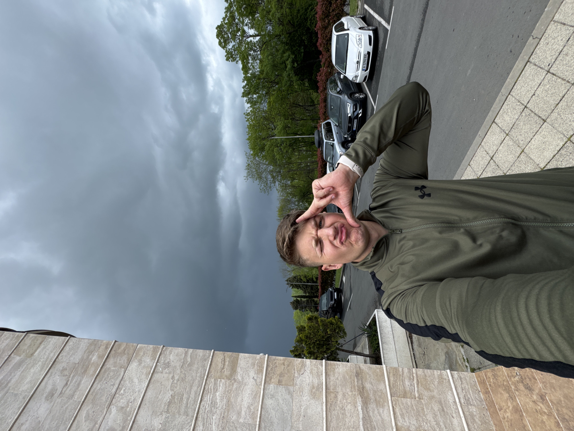 A young man takes a selfie outdoors. He is wearing an olive green Under Armour tracksuit. He is making a humorous face, holding his fingers in a frame shape around his face. Behind him, there is a parking lot with several cars, trees, and an ominous-looking dark gray sky suggesting an approaching storm. The photograph is taken at an angle, with a stone-tiled wall in the foreground.
While the exact location cannot be definitively determined, given the context provided and assuming accuracy, the scene is likely somewhere within the city of Burgas, Bulgaria, though without further information, such as street signs or unique architectural features, specific identification is impossible.