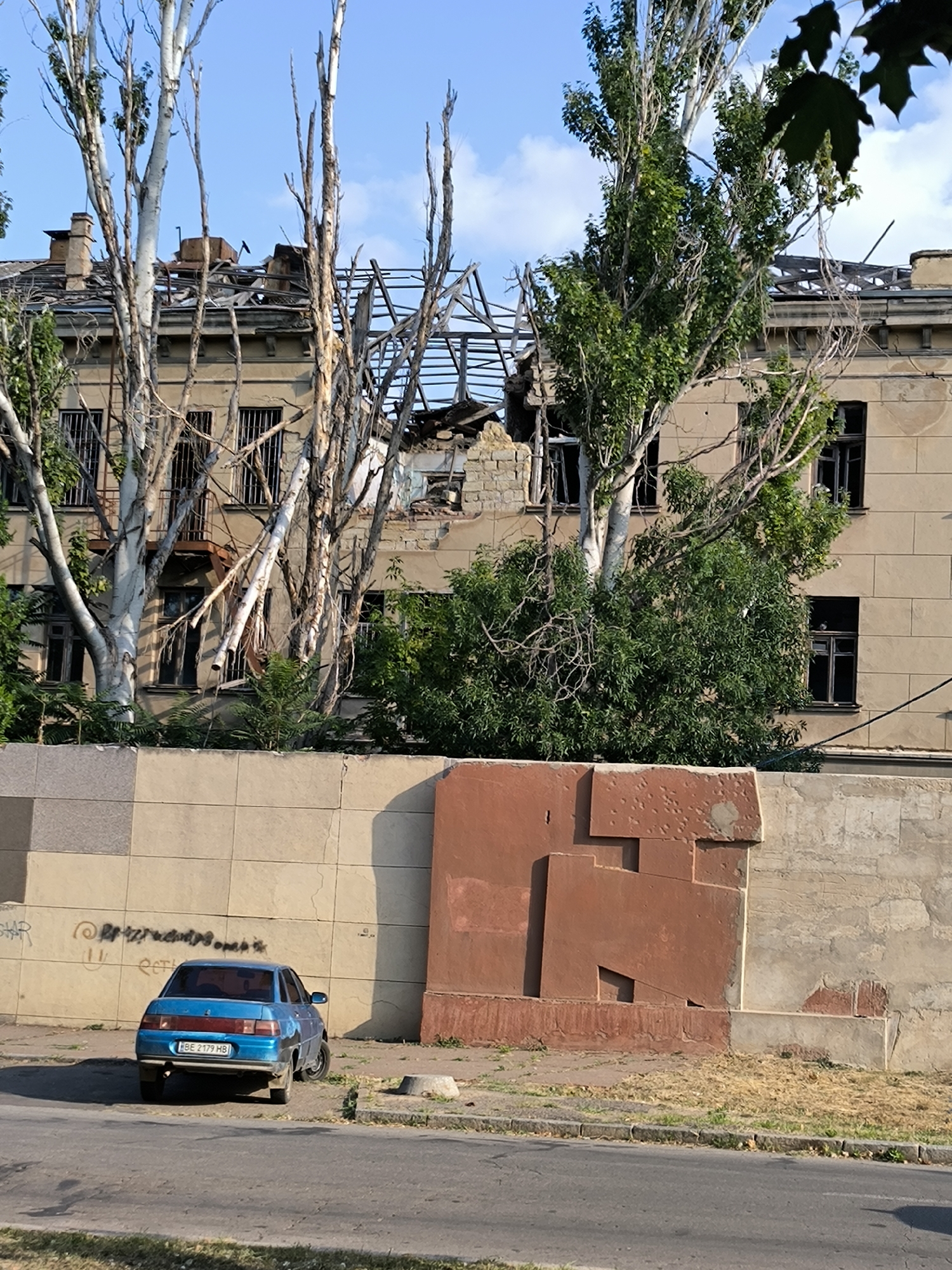 The image shows a destroyed building with a damaged roof and exposed structural elements, possibly due to shelling. Tall, bare trees with some foliage stand in front of the building. In the foreground, a light blue sedan is parked on the side of a paved road, facing away from the camera. A low wall, constructed from large beige rectangular blocks, runs across the middle ground. A large, abstract, rust-colored mural or sculpture is affixed to this wall. The sky is blue with scattered clouds. The scene appears to be an urban environment in Mykolaiv, Ukraine, likely affected by conflict, given the building's condition. There are no people visible in the image.