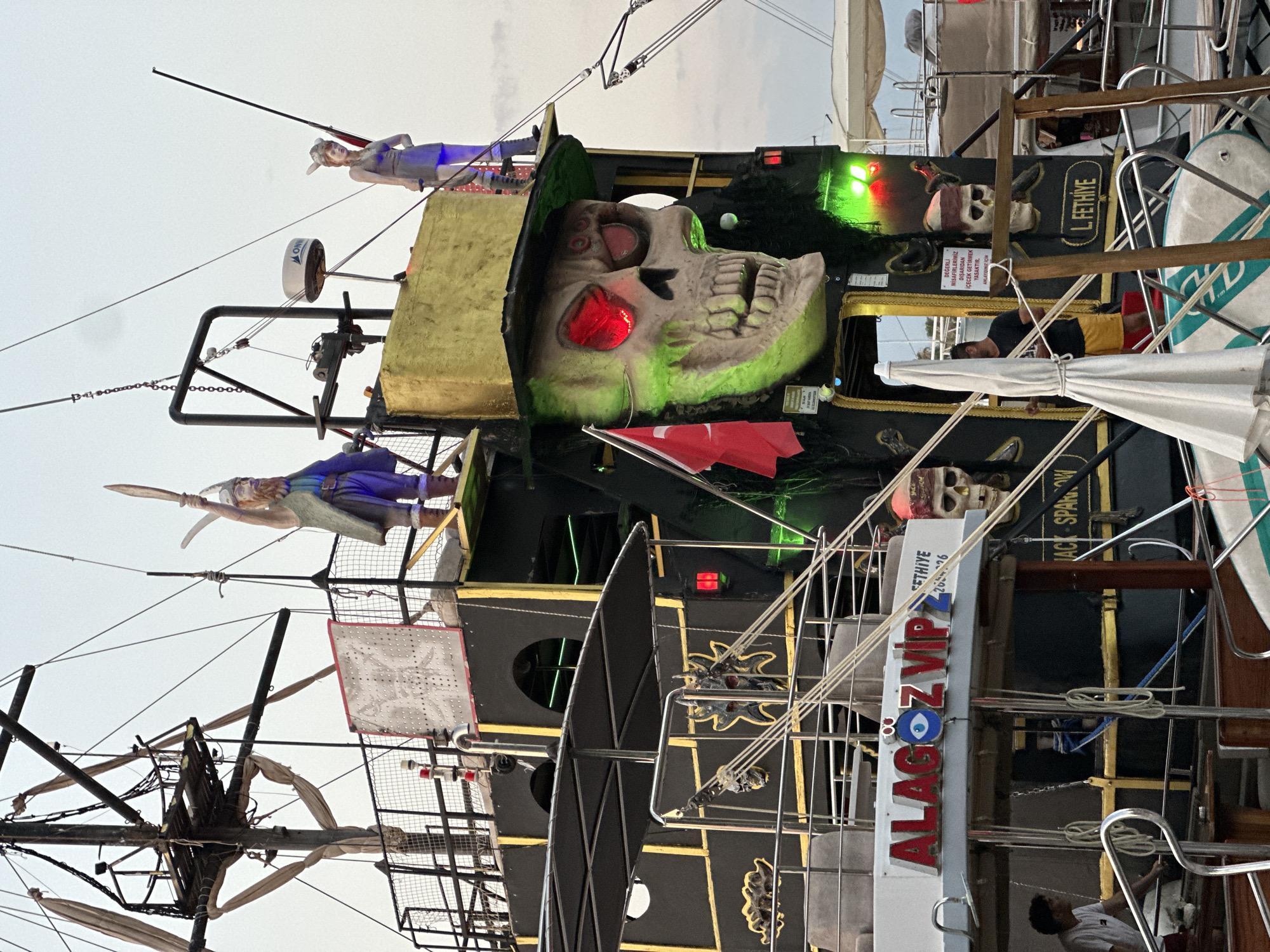 This media file captures a side view of a elaborately decorated boat, likely a tourist vessel, docked in Fethiye, Turkey. The boat is adorned with pirate-themed elements, including a large, glowing skull with red eyes and multiple smaller skull motifs. Several figurines are also present on the boat's upper decks, one appearing to be a pirate or warrior figure.
The boat is painted black with prominent gold accents and lettering. Visible text on the boat includes "ALAGÖZ VIP 2" which appears to be the boat's name, and "L. FETHİYE," indicating its location. Another sign, written in Turkish, states that bringing outside drinks is prohibited. The boat is equipped with masts and rigging, suggesting it is a sailing vessel, though it is currently docked.
The lighting suggests it is either early morning or late afternoon, with the sky appearing overcast. The overall impression is a vibrant and festive atmosphere, characteristic of tourist attractions. The boat's design implies it is used for leisure activities, possibly offering tours or cruises. The presence of a few individuals on the boat, including one person in the foreground, suggests it is operational or being prepared for service.