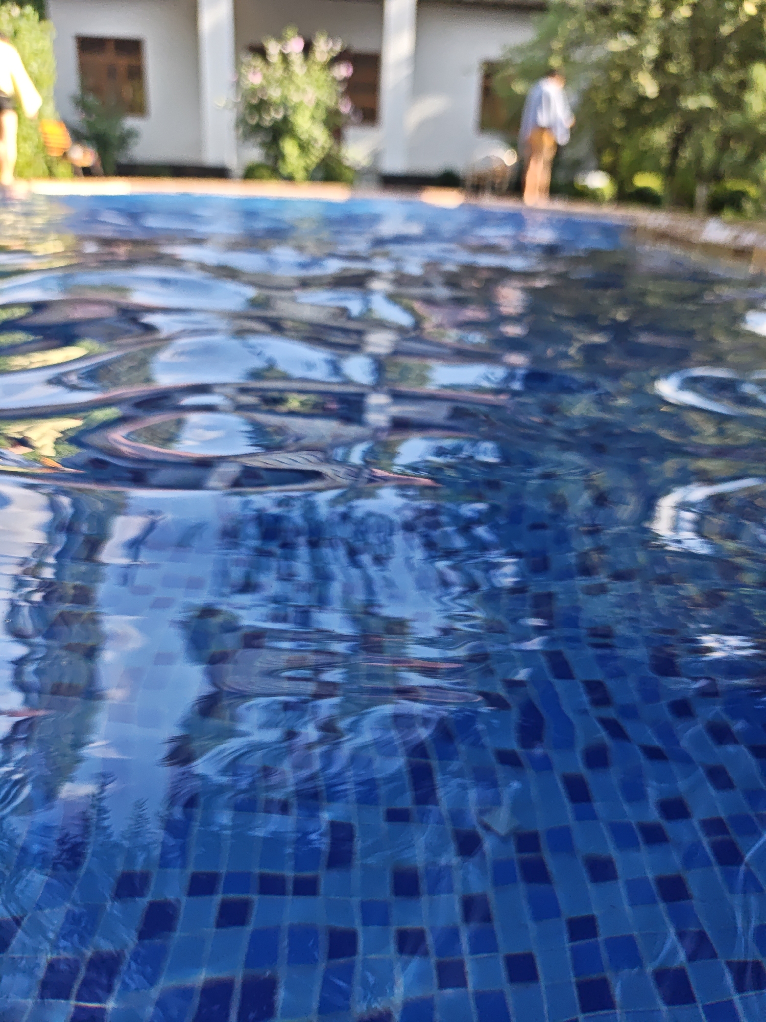 A low-angle, close-up shot captures the rippling surface of a bright blue tiled swimming pool, with the bottom tiles visible through the distorted water. In the blurred background, the facade of a building with windows and lush green trees are discernible, suggesting an outdoor setting likely in Yakkatut, Uzbekistan. On the pool deck, two individuals are faintly visible: one appears to be standing in the water and looking towards the building, while another, further in the background, is walking away from the camera. The overall impression is one of a warm, sunny day at a tranquil location.