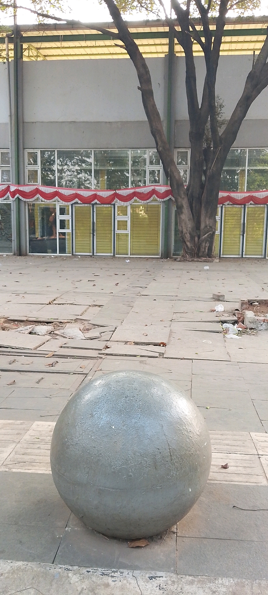 The image shows a large, gray, spherical object made of concrete, situated on a tiled outdoor walkway. The walkway is constructed from large, square tiles, some of which appear to be cracked or displaced, with debris such as leaves and small rocks scattered across the surface. In the background, a building with a light gray facade features a row of large windows with yellow blinds, some of which are open, revealing an interior. A red and white decorative banner is draped across the windows. A large, mature tree with dark bark and bare branches stands prominently in front of the building, its trunk partially obscuring the structure and windows. The area appears to be a public space, possibly near a commercial or institutional building, and the condition of the walkway suggests it might be undergoing maintenance or is in a state of disrepair. This scene is located in Bekasi, Indonesia, as indicated by the specific context of the location.