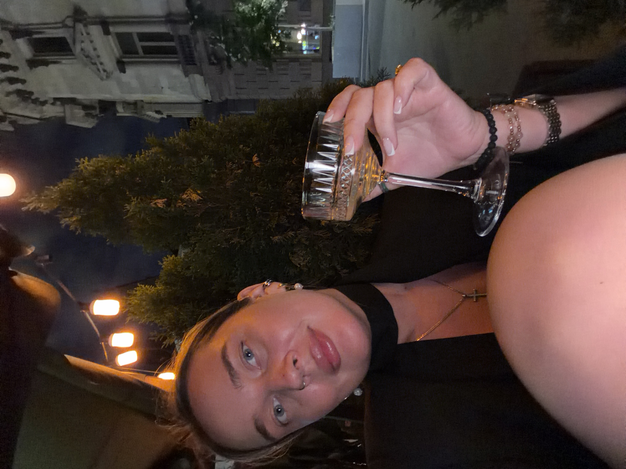 A young woman is seated outdoors at night. She holds a coupe glass in her right hand, wearing multiple bracelets and a watch on that wrist. The woman is wearing a black top, has fair skin, light hair, and blue eyes. She is wearing a necklace with a cross pendant. Behind her is a small evergreen tree and a building, identifiable as being in Moscow, Russia due to the architectural style. Streetlights are visible in the background.