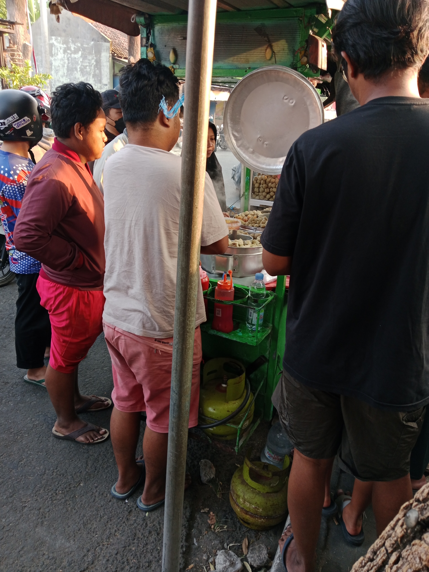 The image depicts an outdoor food stall in Kediri City, Indonesia, possibly on a street. Several individuals are present. In the foreground, a person wearing a maroon long-sleeved shirt and red shorts stands with their hands in their pockets. To their left, another person in a blue and white patterned shirt and black pants is visible from the chest down. To the right of the first person, a person wearing a light-colored long-sleeved shirt and pink shorts is facing away from the camera. Further to the right, a person in a black t-shirt and dark shorts is also seen from behind. A food cart is present, featuring various containers of food, including what appears to be meatballs and other fried items. There are also two green gas tanks, a large silver lid, and bottles of condiments and water on the cart. The ground is paved, and some debris is scattered around the area. The lighting suggests it may be late afternoon or early morning.
