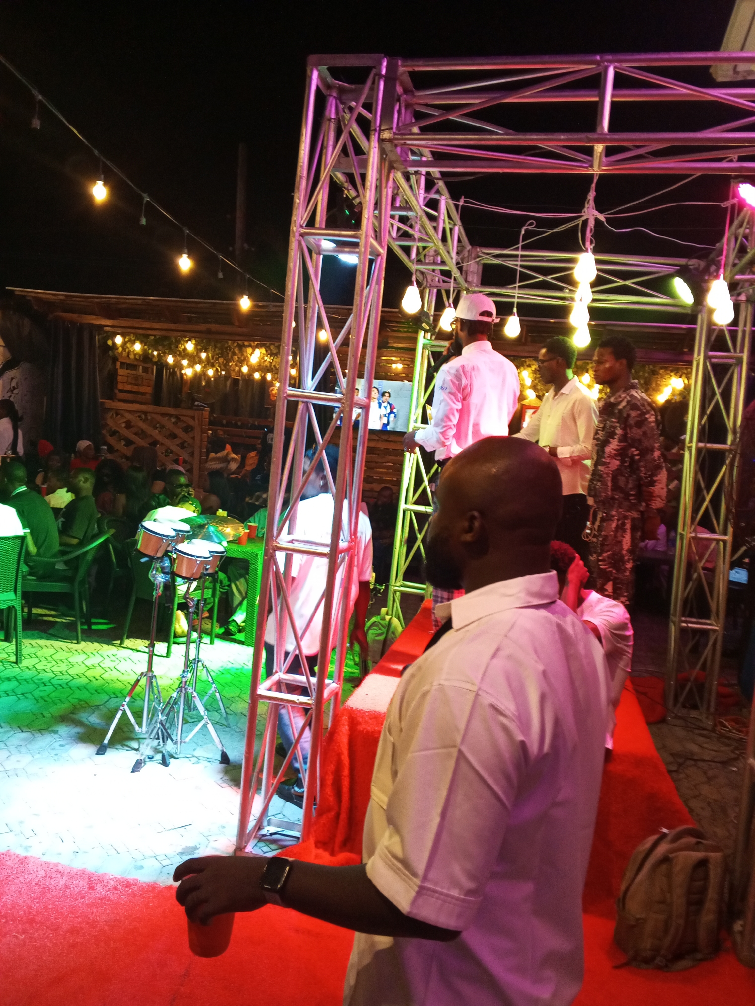 The image captures a lively outdoor event at night, likely a music performance or a party, set in Lokoja, Nigeria. The scene is illuminated by numerous string lights and stage lighting, casting colorful glows of pink and green across the area. A prominent metal stage structure dominates the right side of the frame, adorned with lights and supporting various cables.
Several people are visible, with a focus on a group on or near the stage. One man in the foreground, facing away from the camera, is wearing a light-colored short-sleeved shirt and holding a red cup. Behind him, a man in a white shirt and cap is addressing the crowd, possibly as a presenter or performer. To his right, two other men in light-colored shirts stand, one wearing glasses. Further back, a man in a camouflage patterned outfit is also present.
In the background, an audience is seated at tables, partially obscured by the stage structure and lighting. Some individuals are standing. The ground is covered with a red carpet, with some areas illuminated by green lights. To the left, a drum set with three drums on stands is set up, suggesting a musical performance. The atmosphere appears festive and energetic, typical of a night-time celebration or gathering. The presence of string lights and the outdoor setting indicate it's an evening event. No visible text is present in the image.