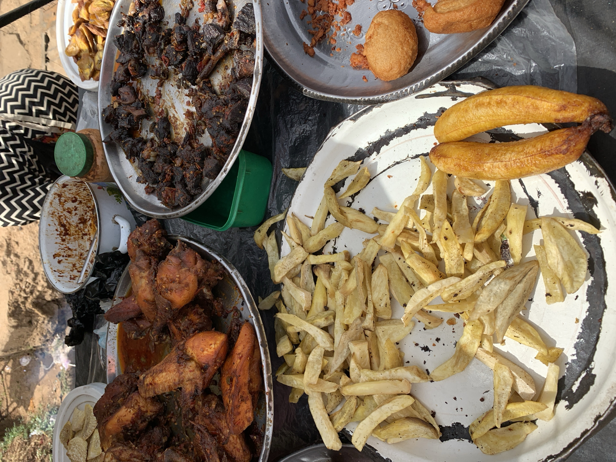 The image depicts several plates of food displayed on a dark surface, presumably for sale. A plate of heavily charred meat sits near a plate with what appears to be fried yam. Another plate contains fried chicken pieces with a reddish sauce. Additionally, there's a plate with fried plantains and what might be puff-puff, a fried dough snack. An enamel pot and some containers are scattered nearby. The overall impression is of street food vendor's setup in an outdoor environment, possibly near New Stadium, Nigeria.