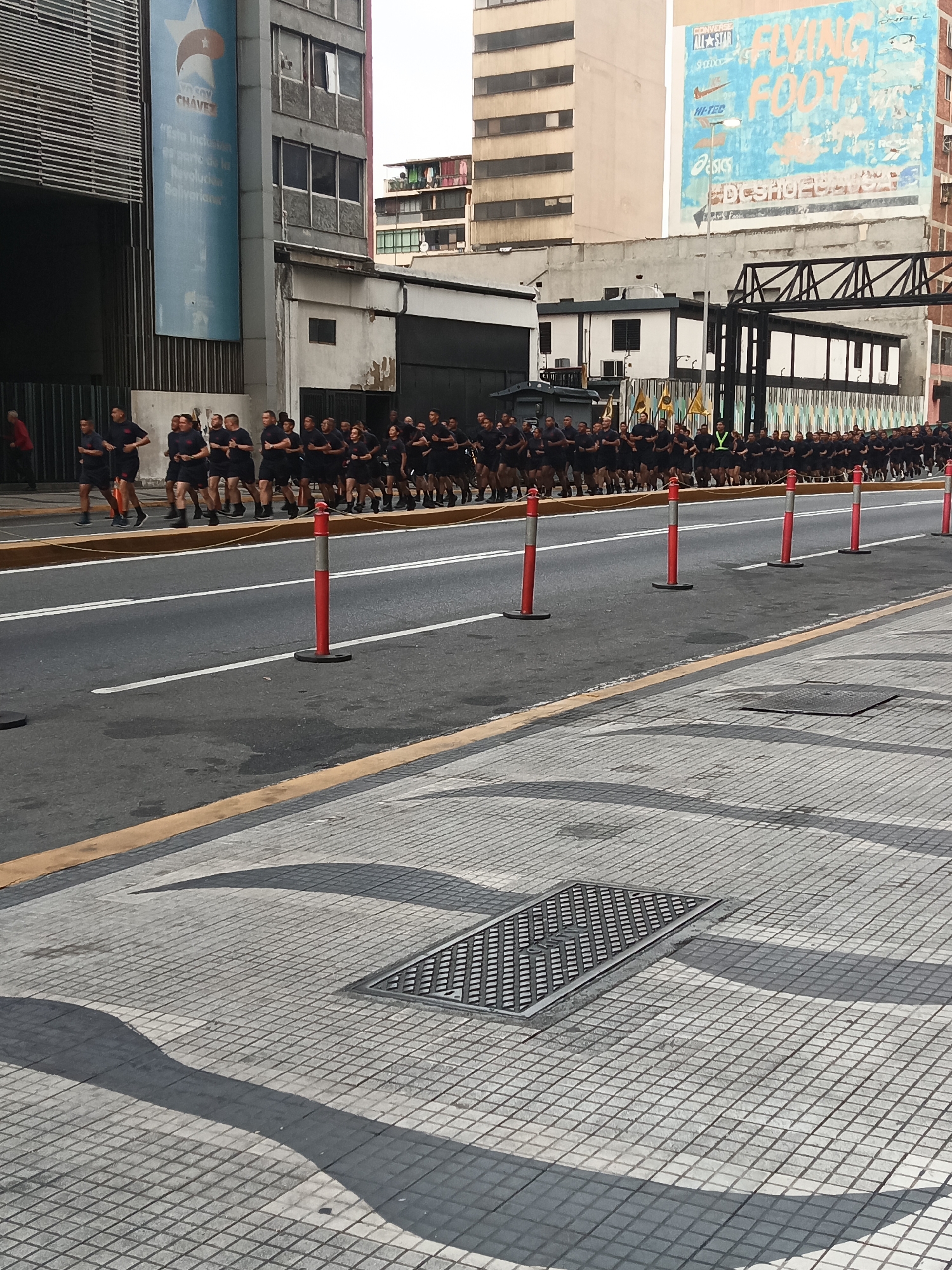 A large group of people in matching dark athletic attire are running on a city street lined with red and white traffic bollards. The road is sectioned off with a yellow curb to the right, where a manhole cover is visible on a patterned concrete sidewalk. In the background, there are tall buildings; one features a large billboard with a stylized star and Spanish text, while another has a faded advertisement with brands like Converse, Nike, and Asics, with the words "FLYING FOOT." The location appears to be in Caracas, Venezuela, given the billboard featuring a star associated with Venezuelan Bolivarianism.