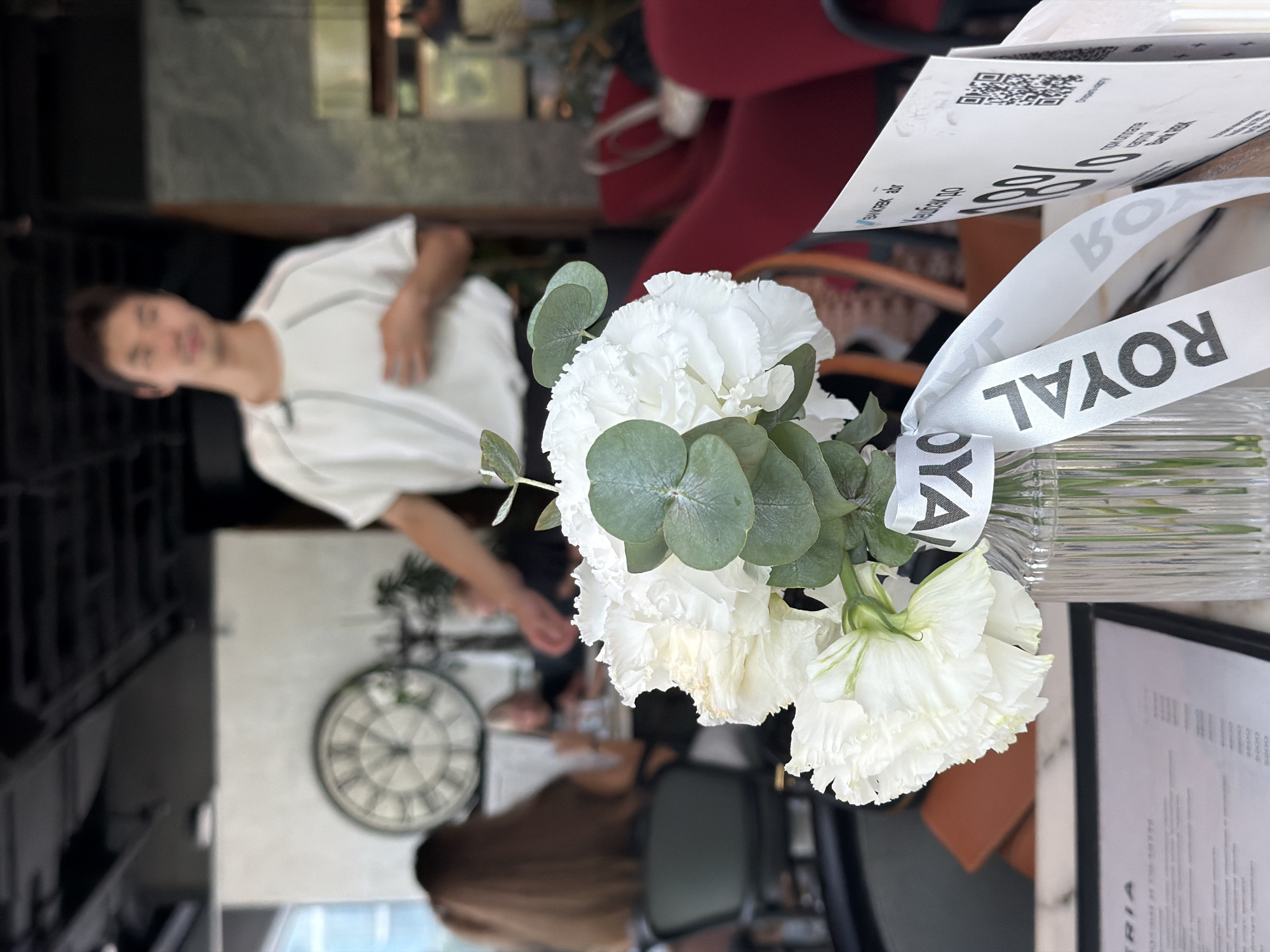 The image depicts a still life scene with a blurred background, set in what appears to be a restaurant or cafe. In the foreground, a glass vase holds a bouquet of white flowers, possibly carnations and eucalyptus leaves, adorned with a white ribbon with the word "ROYAL" printed in black. To the right of the flowers, a promotional card with a QR code and text in Russian is partially visible. The background is out of focus but suggests an indoor environment with patrons seated at tables, a large clock on the wall, and red seating. A person, likely a waiter or staff member, is standing in the background wearing a white shirt. The overall impression is a casually elegant setting, potentially a modern establishment in Almaty, Kazakhstan, given the presence of Russian text on the promotional material.