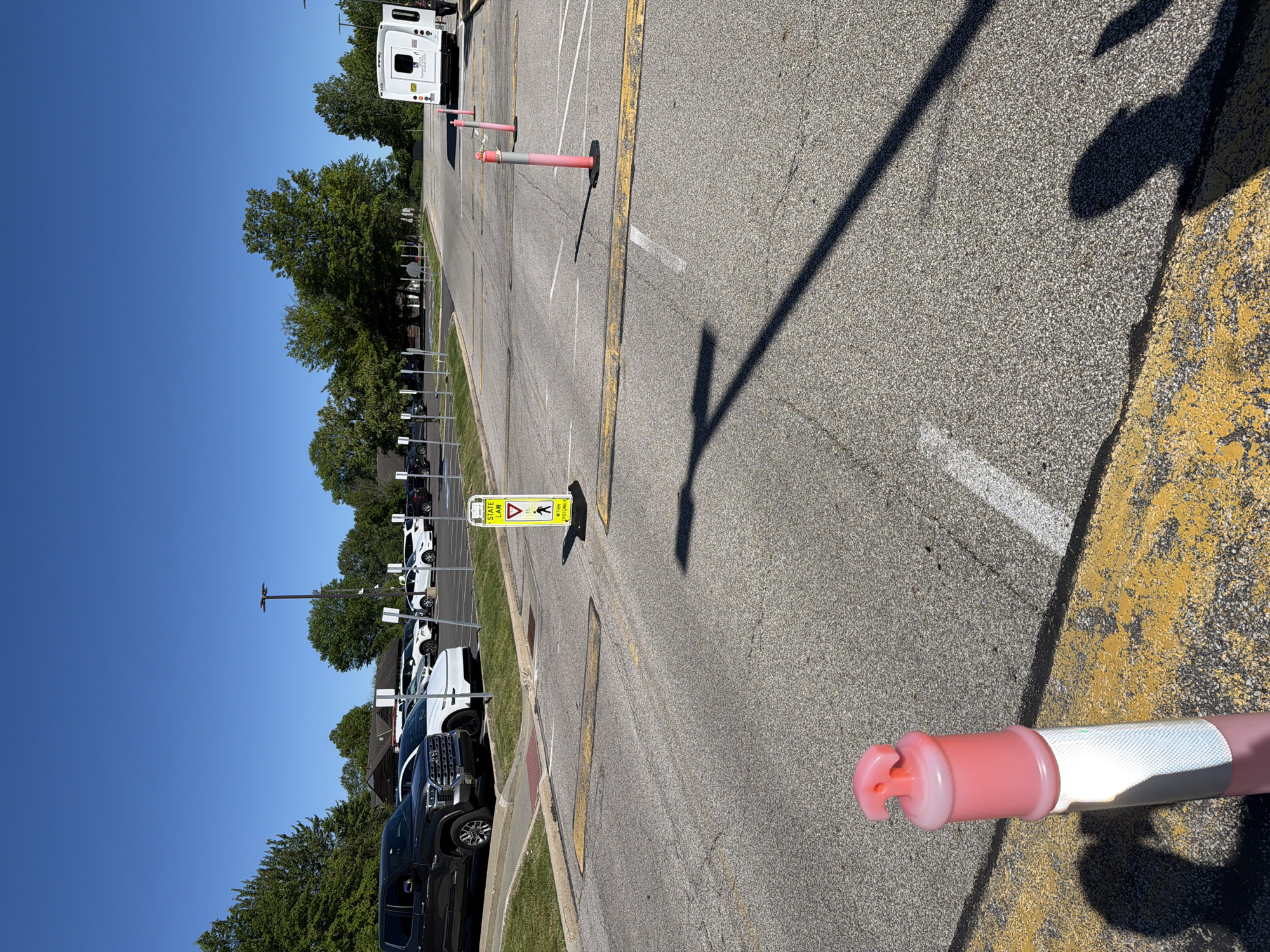 This outdoor image depicts a paved parking area in Beachwood, United States of America, bathed in bright sunlight under a clear blue sky. The scene suggests a daytime setting, likely mid-morning or afternoon, given the length and angle of the shadows cast across the asphalt.
In the immediate foreground on the right, a pink and white reflective safety bollard lies partially visible. Spaced along the midground of the paved surface are several upright safety bollards, colored pink and gray, which appear to mark lanes or direct traffic flow. The asphalt itself is marked with yellow and white lines, typical of a parking lot or road.
A prominent yellow sign stands on a metal post in the middle distance. This sign features a triangular yield symbol, an arrow pointing to a pedestrian crossing icon, and includes the text: "STATE LAW", "YIELD TO", and "WITHIN CROSSWALK", indicating a pedestrian right-of-way area.
To the left of the paved section, a row of vehicles, including dark-colored pickup trucks and white cars, are parked. These vehicles are situated alongside a grassy strip and a line of verdant trees and bushes. In the background on the far right, a white utility or service truck with a large, boxy rear compartment is visible, parked among more trees. The overall impression is a functional outdoor space emphasizing safety and traffic organization. No people are visible, and the scene is static.