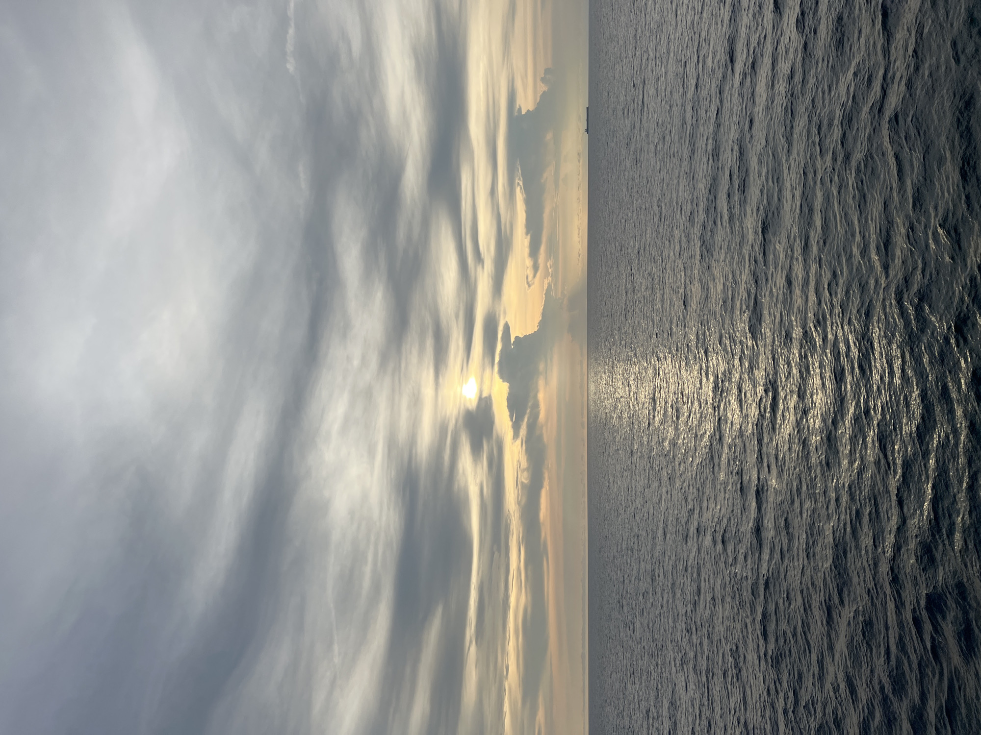 The image depicts a wide expanse of the sea under a cloudy sky. The sun is partially obscured by clouds, casting a golden glow on the horizon and creating a shimmering reflection on the water's surface. The sea is a deep blue-gray with small waves, indicating a moderate chop. In the distance, a lone cargo ship is visible on the right side of the frame, a small silhouette against the vastness of the ocean and sky. The clouds are a mix of gray and white, with some appearing wispy and others more solid, suggesting varied atmospheric conditions. The overall mood is serene and expansive, characteristic of an open sea view. There are no people or urban structures visible. The location is identified as Unknown, Unknown, indicating a lack of specific geographical data.