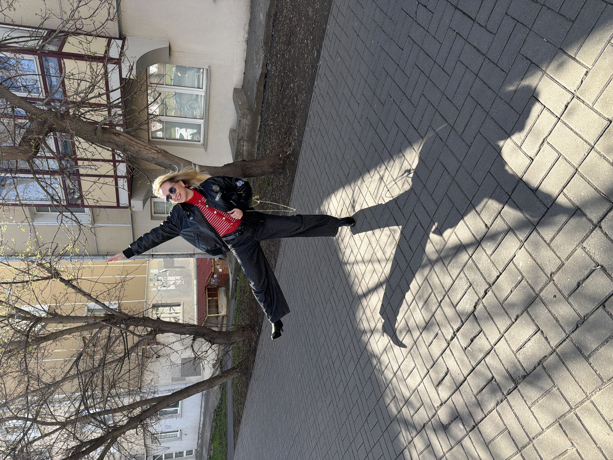 The image, oriented correctly, captures a blonde woman in a dynamic pose on a paved sidewalk on a sunny day. She appears to be in her 30s or 40s, dressed in a black leather jacket, a red and white striped top, dark trousers, and black heeled boots. Sunglasses are perched on her face, and she wears red earrings. Her hair is flowing, suggesting motion, as she smiles and extends one leg high into the air with her arms outstretched, as if jumping or kicking. A small black backpack is visible on her back.
Long, distinct shadows are cast on the light grey herringbone-patterned sidewalk, indicating the sun is low in the sky, likely morning or late afternoon. In the background, multi-story residential buildings with numerous windows line the street. Several trees with bare branches and hints of new green buds signify springtime. A narrow strip of soil and sparse vegetation separates the sidewalk from the buildings. The urban setting is consistent with the provided location context of Izhevsk, Russia. A small white logo resembling "DKNY" is visible on the left arm of her black jacket. The overall mood is joyful and energetic.