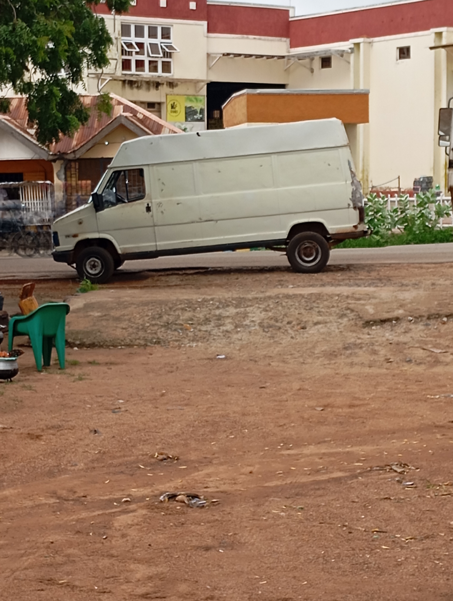 A light-colored, boxy van is parked on a dirt road in Shendam, Nigeria. The van has a high roof and appears to be an older model. To the left of the van, a bright green plastic chair is positioned next to a dark pot or container. In the background, there is a building with a red roof and cream-colored walls, and a section of a building with a rusty corrugated metal roof. The ground is covered in reddish-brown dirt with some scattered debris. A few trees with green foliage are visible on the left side of the image. The sky is overcast.