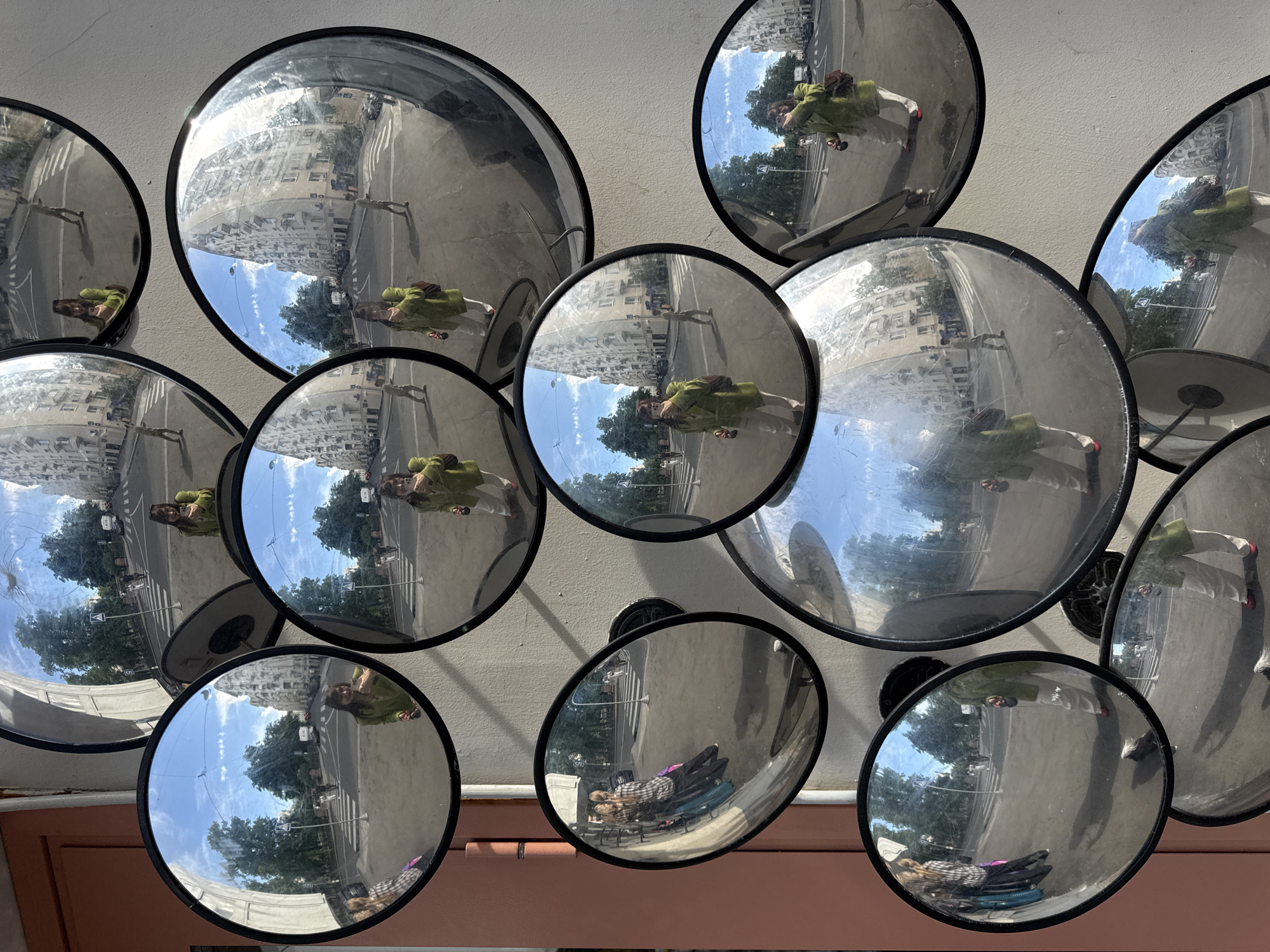 A collection of convex mirrors of various sizes are mounted on a light-colored wall, creating a fragmented and distorted view of an urban scene. The primary subject reflected in multiple mirrors is the photographer, seen from a high angle, wearing a light green jacket or coat, light-colored pants, and red shoes or sandals, holding a phone.
The reflections within the mirrors collectively depict a bright, sunny day in a city, consistent with Moscow, Russia. The sky is blue with scattered white clouds, and large, green deciduous trees line what appears to be an asphalt street. Distinct white crosswalk markings are visible on the road. Tall, light-colored residential or commercial buildings with numerous windows are prominent in the background of many reflections.
Beyond the photographer, other people are subtly reflected in the street scenes, appearing to walk or stand. Notably, one of the lower mirrors captures a reflection of a person lying down, possibly on a bench, covered with a plaid blanket. The scene is illuminated by bright daylight, suggesting midday or early afternoon. The overall effect is a multi-perspective, surreal portrayal of a vibrant city environment under clear weather. No discernible text is visible in the image.