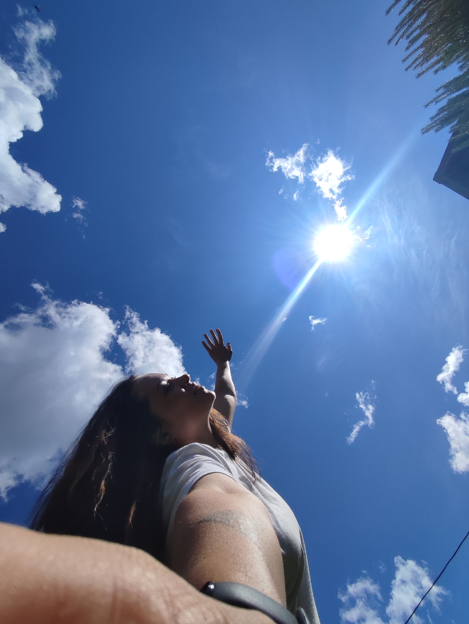 A low-angle outdoor photograph depicts an individual with long, dark hair, wearing a white shirt, positioned with their left arm and shoulder in the immediate foreground. The person's face is shown in profile, looking upward towards a brightly shining sun in the upper right quadrant of the image. Their right arm is raised, hand open, extending towards the sun. A black wristband or watch is visible on the left wrist. The sky is a vibrant blue with scattered white cumulus clouds, more concentrated on the left side of the frame. Strong sun rays and lens flare emanate from the sun. In the upper right corner, dark green conifer branches and a dark angular structure, possibly a building, are partially visible. A thin dark line, resembling a utility wire, traverses the lower right section of the sky. The scene is situated in the city of Фоки, Russia.
