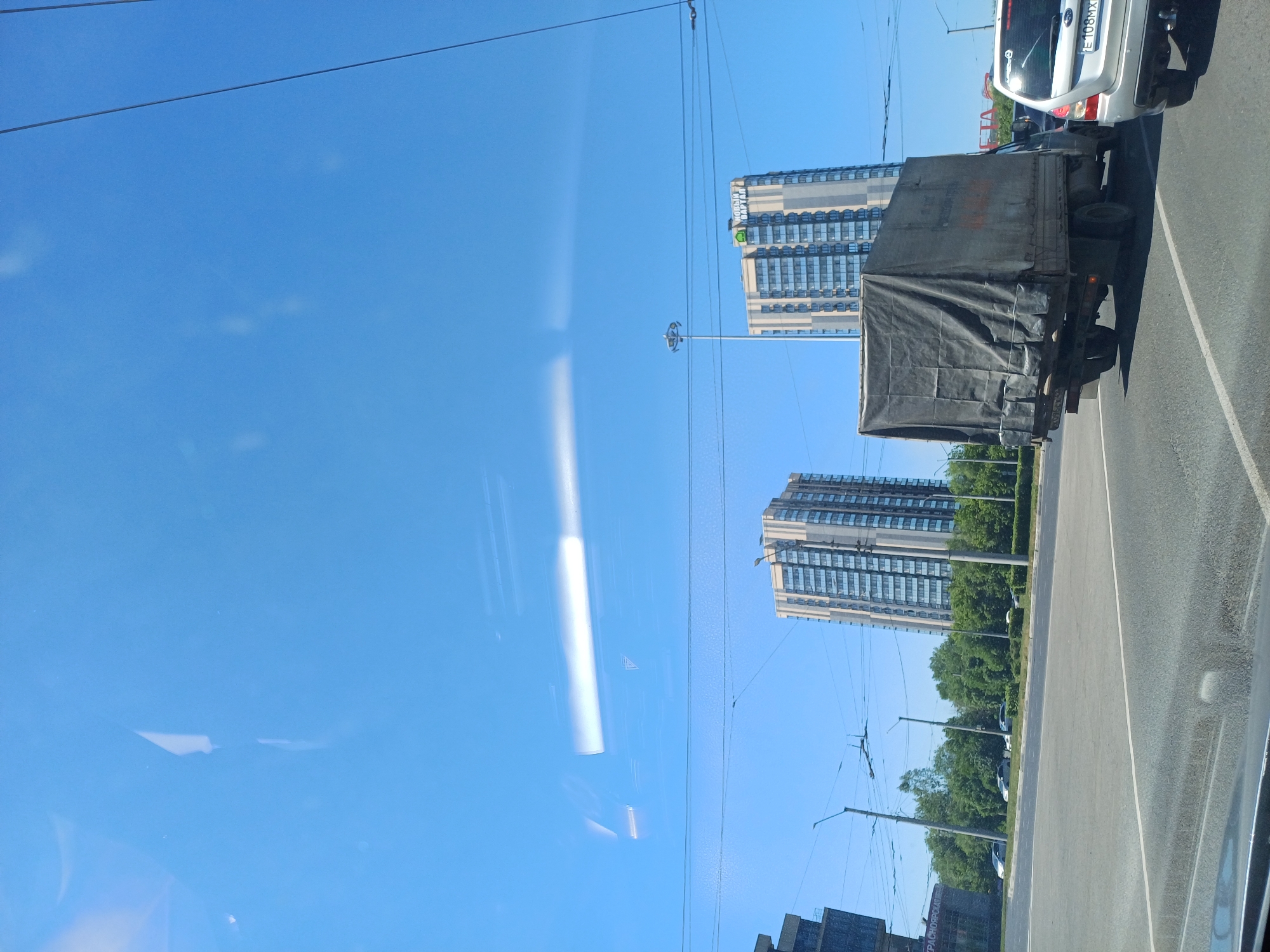 A street-level, eye-level outdoor medium shot captures a sunny day in a city. A large, dark gray tarp-covered truck, with visible tire tracks on its dirty surface, is the central focus, driving away from the camera in the foreground. To the right, the rear of a silver car is visible, its license plate reading "E 108MX". In the background, two tall, modern residential buildings with multiple balconies are prominently featured against a clear blue sky. Trees line the median between the road and the buildings, and power lines crisscross the scene. A pedestrian signal light is visible near the center of the image. The overall environment suggests a typical urban setting on a bright day.
