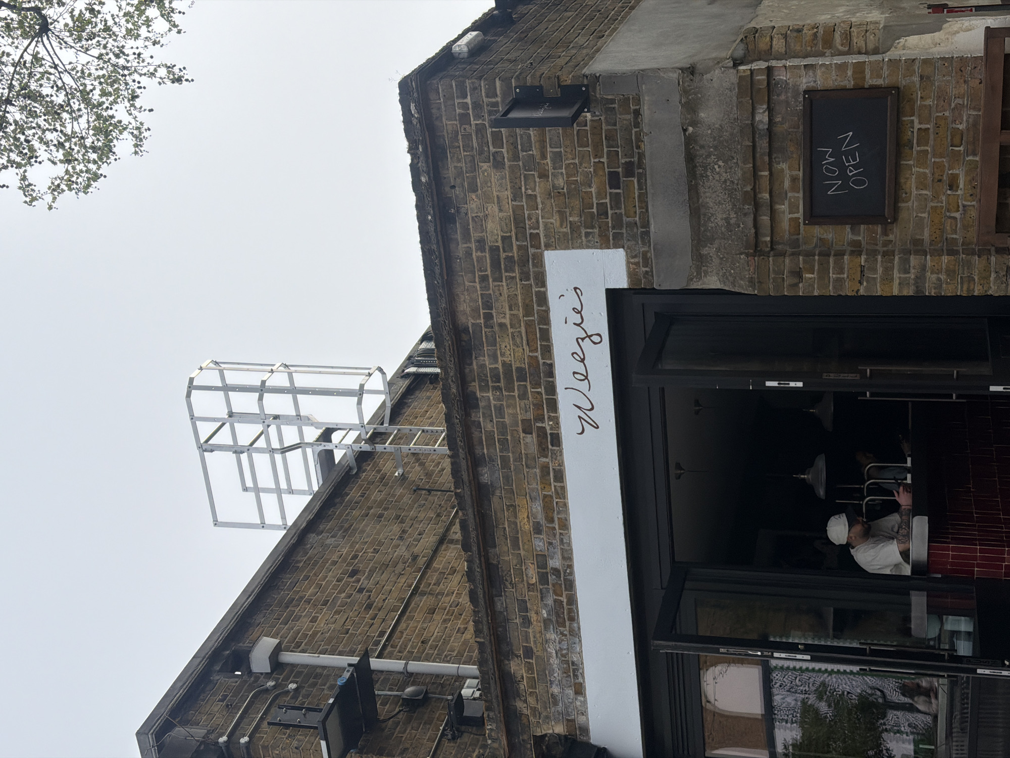 A ground-level, upward-angled shot captures the facade of "Weezie's," a restaurant in London, United Kingdom. The restaurant is housed in a brick building, with a white sign displaying the restaurant's name vertically. A "Now Open" sign hangs on the brick wall beside the black-framed entrance.
Inside, a man wearing a white cap and shirt is visible. A complex metal framework is installed on the roof of the building. The sky is overcast, and tree branches are visible in the upper left corner of the frame.
