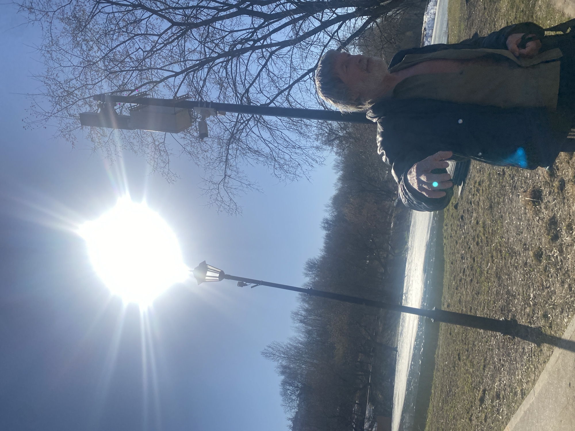 A man stands outside on a sunny day, facing away from the camera, with his hand outstretched. A lamppost is positioned to his left. The area appears to be a park or outdoor space with trees in the background. Based on the style of the lamppost and the appearance of the environment, the location is likely in Moscow, Russia.