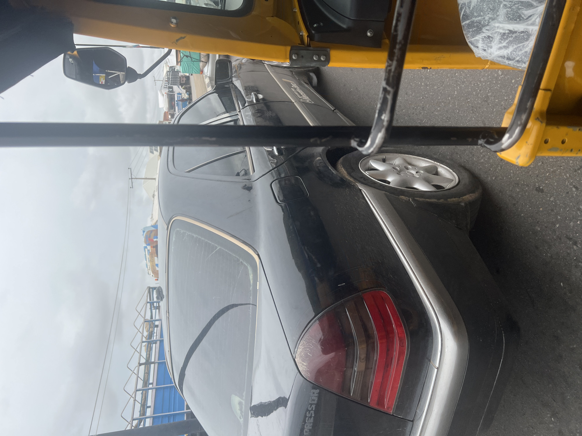 The image captures a traffic scene in Jalingo, Nigeria, with a black Mercedes-Benz sedan prominently featured in the foreground, seen from its rear passenger side. The car appears dusty, with visible water streaks on its rear window, suggesting recent rain or exposure to the elements. The taillights are partially smoked. A silver trim runs along the lower body of the car, and the rear bumper is a dark grey. The wheels are silver alloy with a multi-spoke design. The car's badging on the trunk area appears to read "COMPRESSOR" and possibly "C320".
To the right of the Mercedes, a portion of a bright yellow motorized rickshaw, also known as an "Okada" in Nigeria, is visible. Its metallic frame and yellow bodywork are distinct. The rickshaw's interior, partially covered with a clear plastic sheet, can be seen.
In the background, other vehicles are present, including trucks with blue and green cargo covers, and a white truck with a metal grate structure. Power lines and utility poles are visible against an overcast sky, typical of a cloudy day. The road surface is asphalt. The overall scene suggests moderate traffic in an urban or semi-urban setting.
