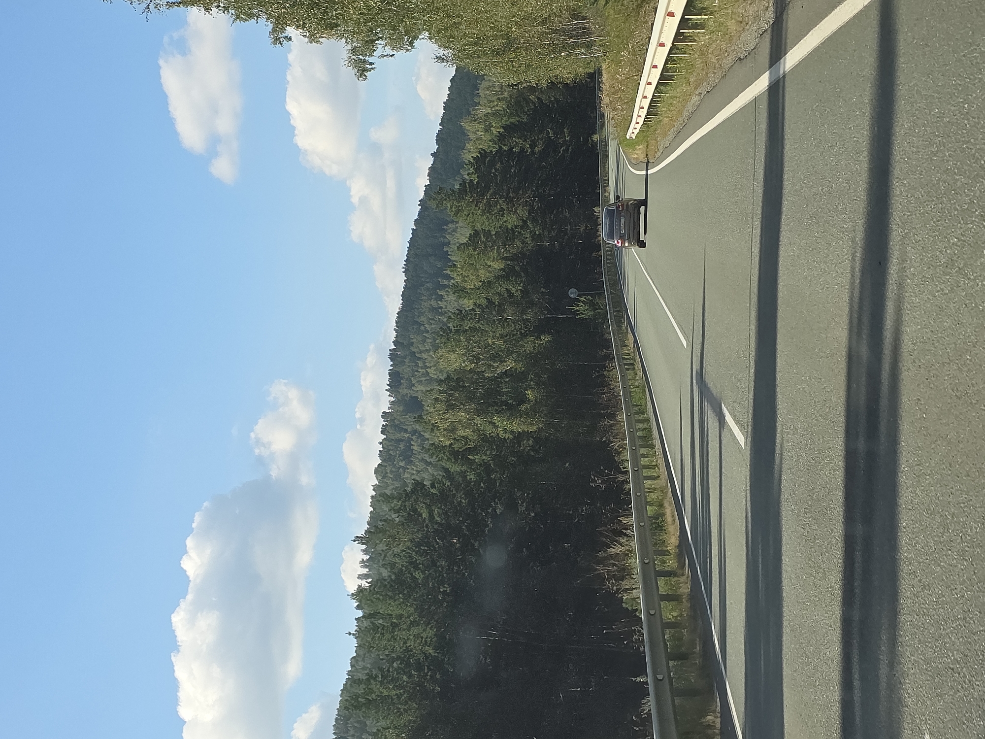 A dark-colored sedan is driving on a paved road through a wooded area with a hill in the background. The car is in the right lane, and there is a white line marking the edge of the lane. The road has a light gray asphalt surface and is lined with guardrails. The surrounding area is covered with green trees, and the sky is blue with some white clouds. The image appears to have been taken from another vehicle driving behind the sedan. The location is identified as Saktaevo, Russia.