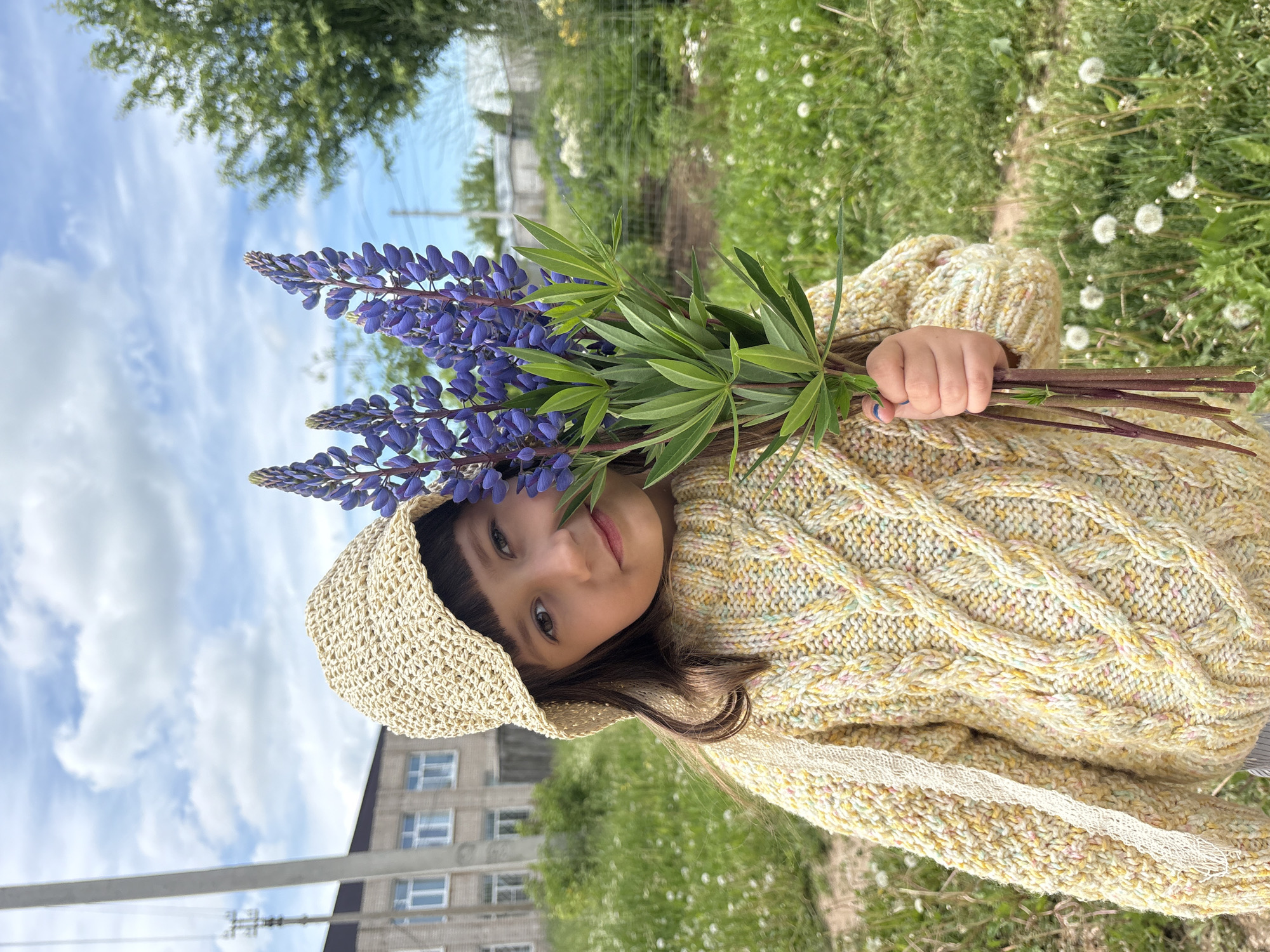 This image features a young child, likely a girl, holding a bouquet of purple lupine flowers. The child has dark hair with bangs, is wearing a light yellow or cream-colored cable-knit sweater with subtle multicolored specks, and a straw hat. She looks at the camera with a gentle, slightly smiling expression, conveying a sense of innocence and happiness. Her hand, with light blue nail polish on some fingers, securely holds the stems of the flowers.
The setting is outdoors on a bright day, under a clear blue sky with white, fluffy clouds. In the background, there is a lush green grassy area dotted with white dandelion seed heads. Further back, green trees are visible, along with a multi-story building, possibly an apartment complex, featuring numerous windows. Some faint utility lines or structures are also present against the sky. The overall scene suggests a pleasant day, likely late morning or afternoon, spent in a natural or semi-urban environment. The location context provided is Фоки, Russia. There is no visible text in the image.