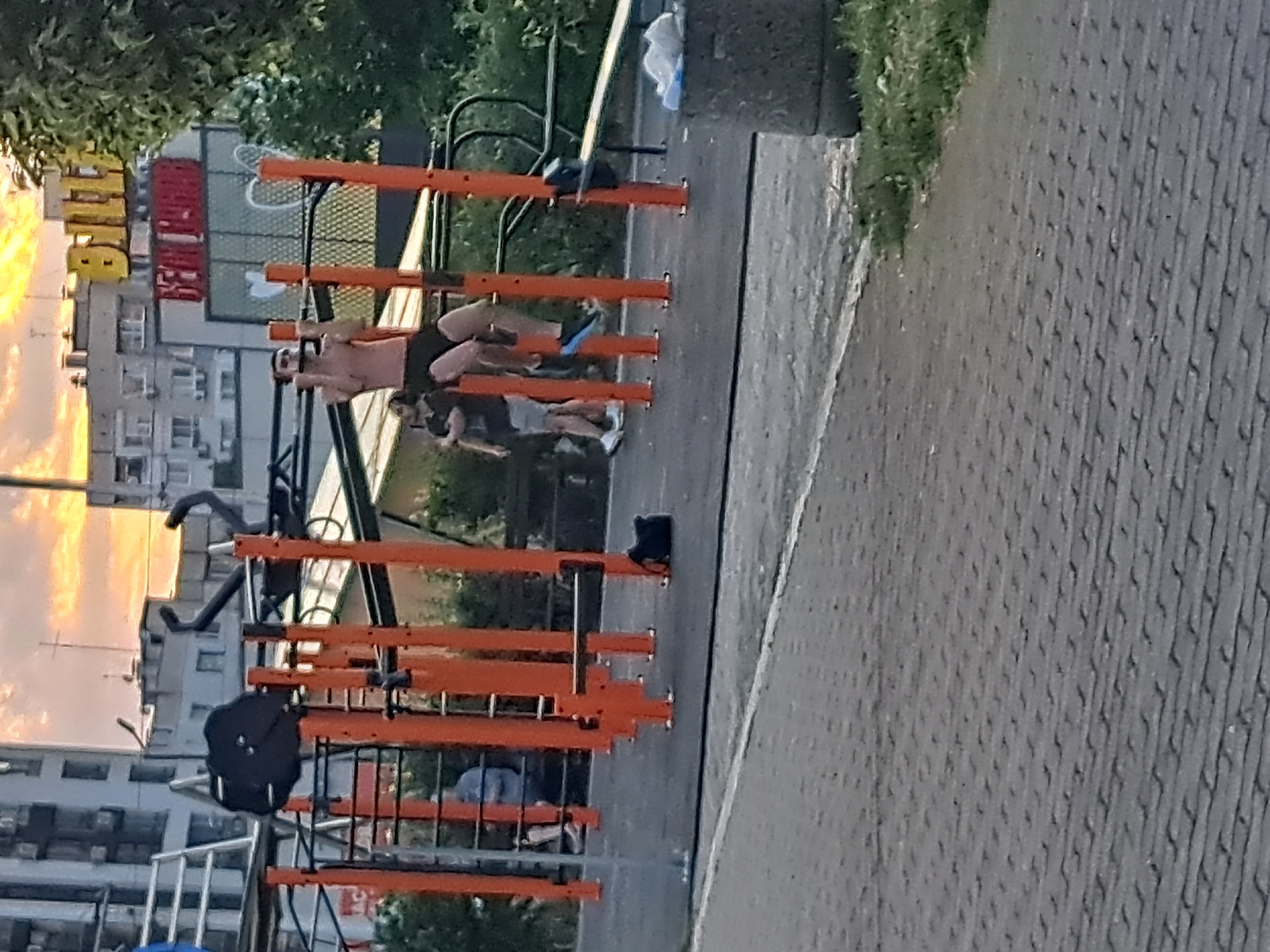 This media file shows an outdoor exercise area in Sofia, Bulgaria. The scene features several people using orange and black exercise equipment. One person is lying on their back on the equipment, with their legs extended upwards. Another person is in a seated position, appearing to rest. The equipment itself consists of bars and platforms, suggesting it's designed for various calisthenics exercises.
The background shows apartment buildings and trees, indicating an urban environment. The lighting suggests it might be late afternoon or early evening, with the sun casting a warm glow on the buildings. The ground is paved with what appears to be patterned concrete or rubberized surfacing, common in public recreational areas. Some of the apartment buildings have visible windows, and one building has yellow and red signage. There is no visible text that is clearly legible. The overall impression is of a public park or community space where people are engaged in physical activity.