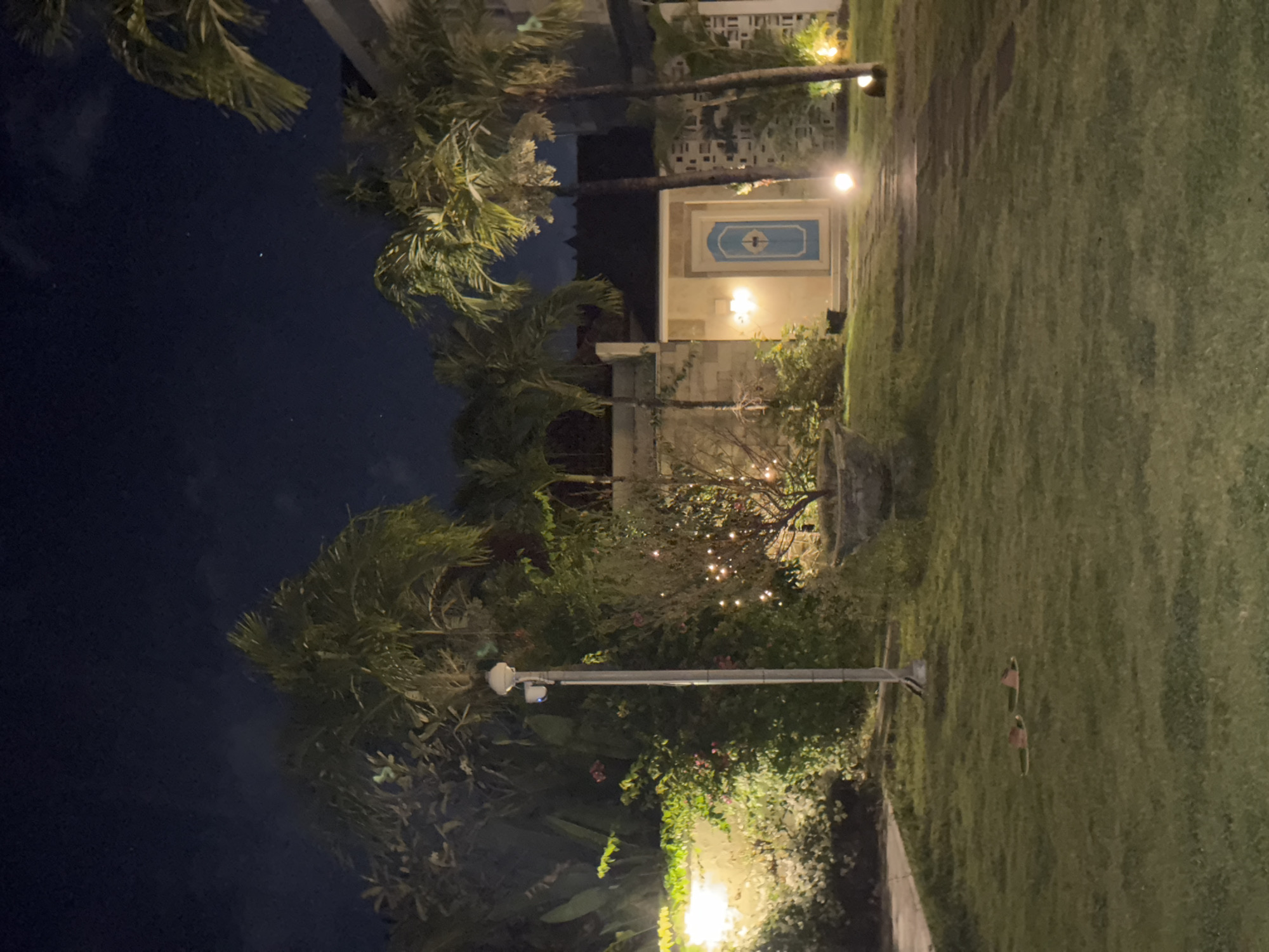 A nighttime view of a well-lit garden in Kuta Selatan, Indonesia, shows a paved pathway leading to a house with a light blue door. Palm trees and lush vegetation surround the area, with string lights illuminating a small tree. A lamppost with two security cameras is positioned in the foreground, and a pair of flip-flops rests on the grass. The sky is dark with a few visible stars.