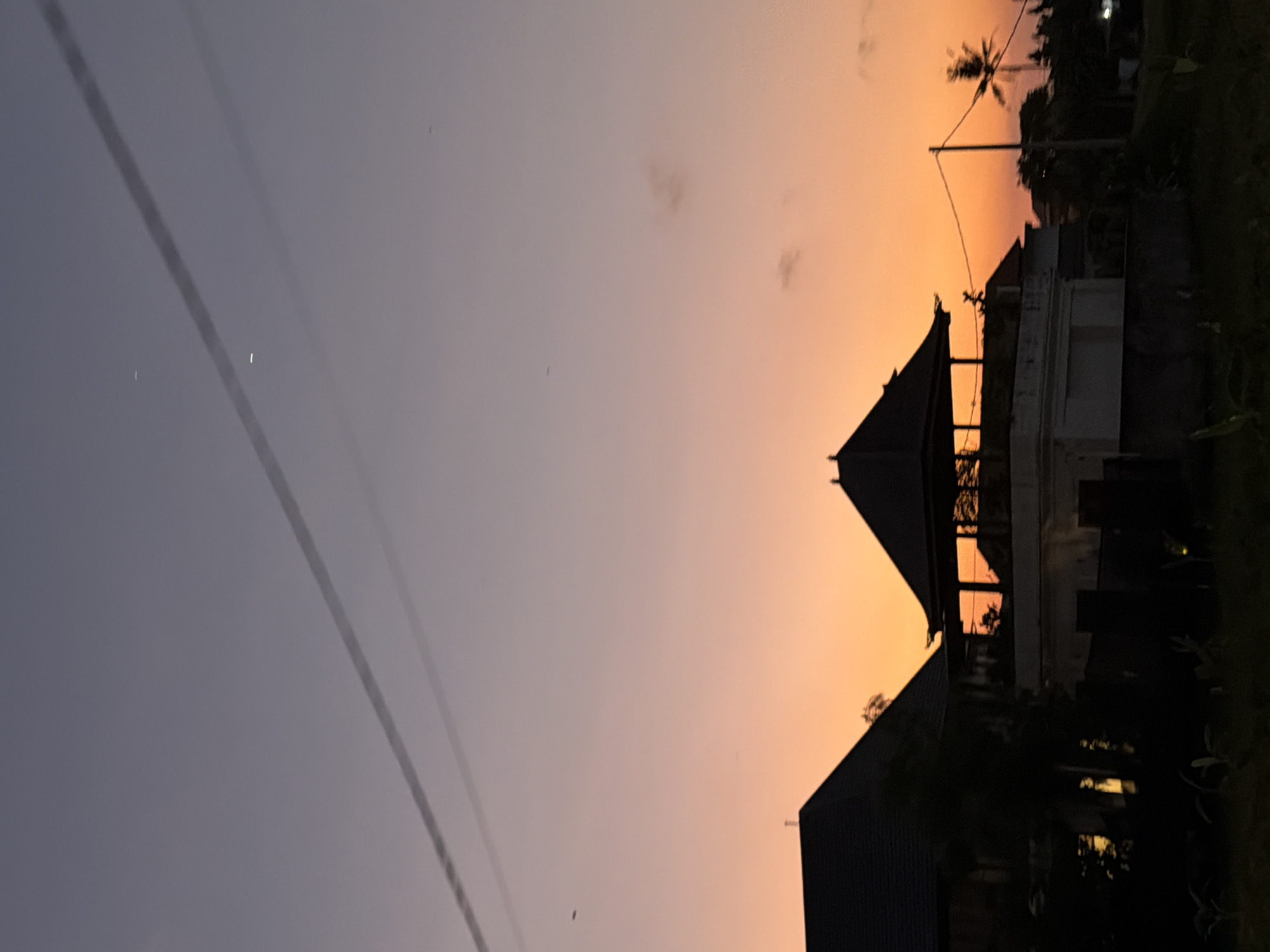 An evening scene in North Kuta, Indonesia, depicts a sunset or dusk transition with a gradient sky shifting from a deep purplish-grey at the zenith to vibrant orange and yellow hues near the horizon. Two distinct, dark, parallel power lines traverse the upper left quadrant of the sky. Two small, luminous white points, possibly celestial bodies or distant artificial lights, are visible in the upper mid-right. Sparse, dark specks, likely birds or insects, are scattered across the sky, along with a few faint, wispy clouds in the mid-right.
The foreground is dominated by the dark silhouettes of architectural structures and vegetation. On the left, a building with a dark, steeply pitched roof is partially obscured by dense, dark foliage. Centrally, a traditional Balinese-style open pavilion structure with a multi-tiered dark roof stands elevated on a lighter-colored base, possibly a light-toned wall or building. This central structure exhibits an internal orange glow, possibly from artificial lighting or reflected ambient light. To the right, another dark building with a pitched roof is visible, along with a solitary, tall palm tree silhouetted against the sky. A utility pole with overhead wires is present near the palm tree, and a faint, small white light source is discernible further right. The immediate foreground consists of dark, indistinct ground with faint outlines of low-lying vegetation. No human figures are present.