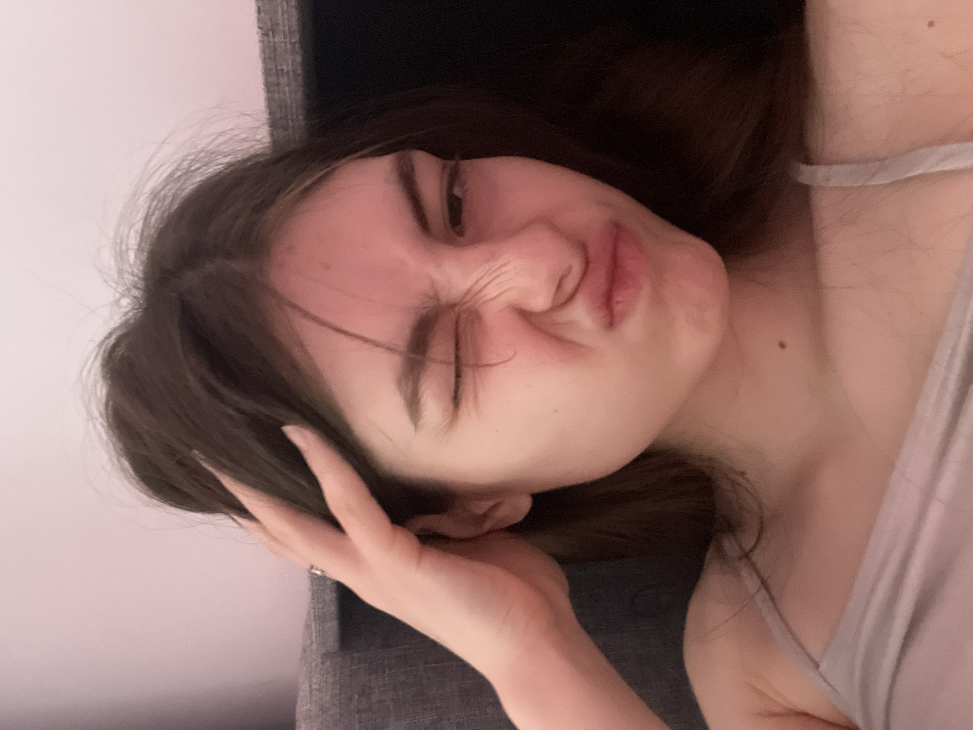 A young woman with brown hair is making a facial expression, wrinkling her nose and squinting one eye. Her hand is near her head. She is wearing a gray tank top and is sitting on or against a gray, upholstered surface. The background is plain and out of focus. Location: Unknown, not Saint Petersburg, Russia.