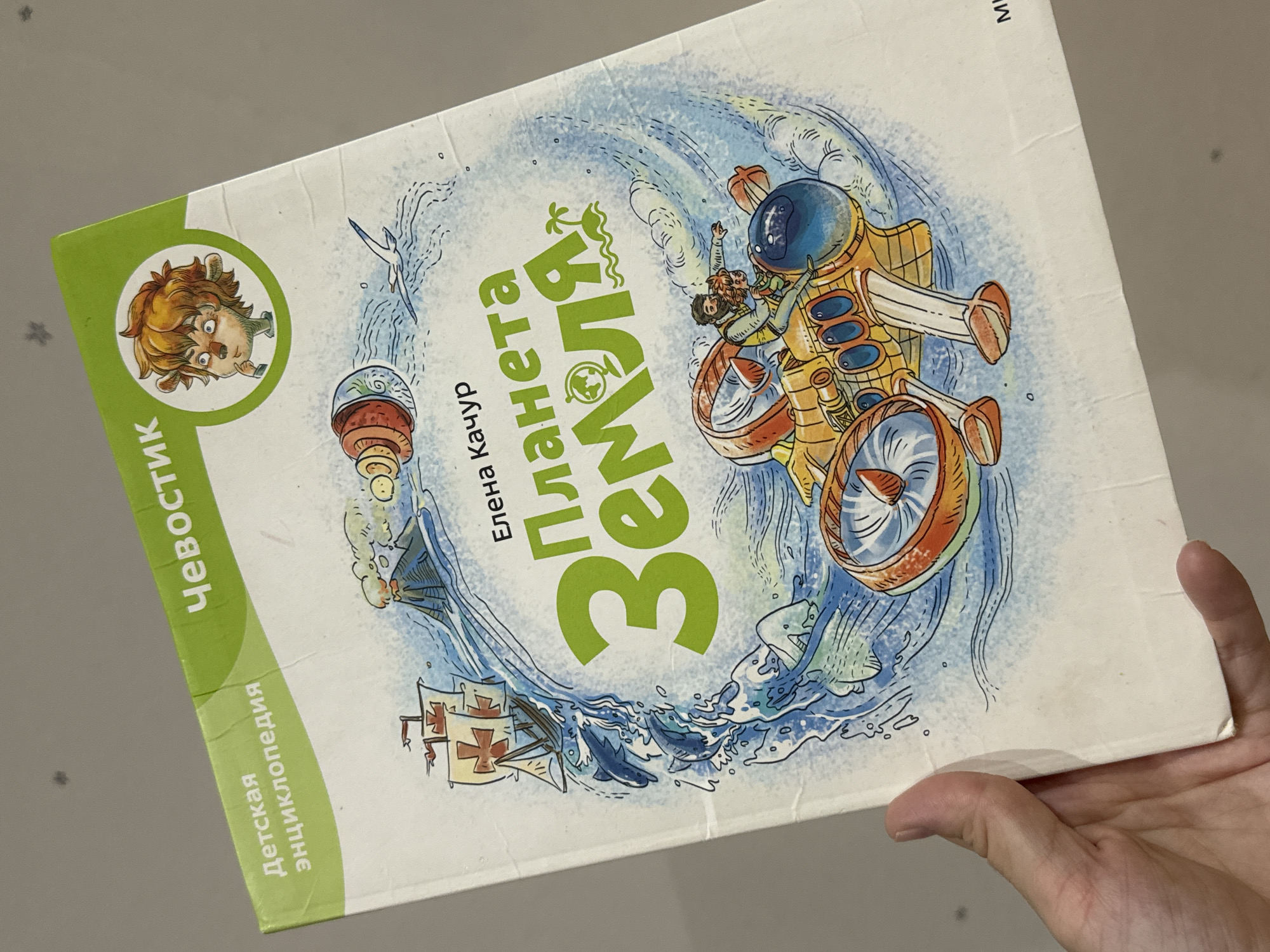 A hand is holding a children's encyclopedia titled "Planet Earth" in Russian. The book cover features cartoon illustrations of various modes of transport and sea creatures. The location is not identifiable as Chaikovsky, Russia from the given image.