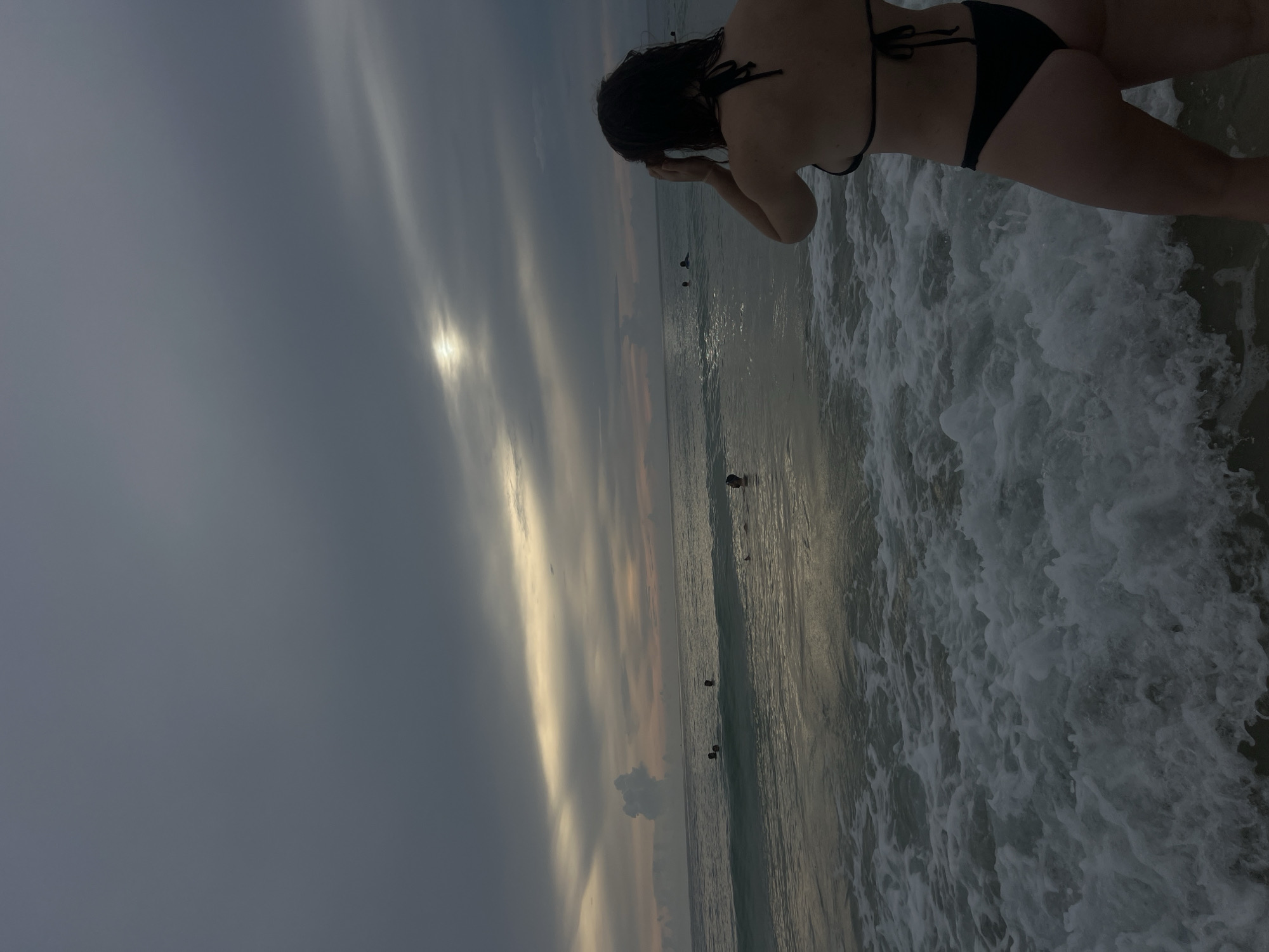 A woman in a black bikini stands at the edge of the ocean, facing away from the camera. The sky is overcast with a sliver of sunlight breaking through the clouds, creating a dramatic effect. The ocean is choppy with waves crashing on the shore. Several other people are visible in the water in the distance. The location is a beach in Choeng Thale, Thailand.