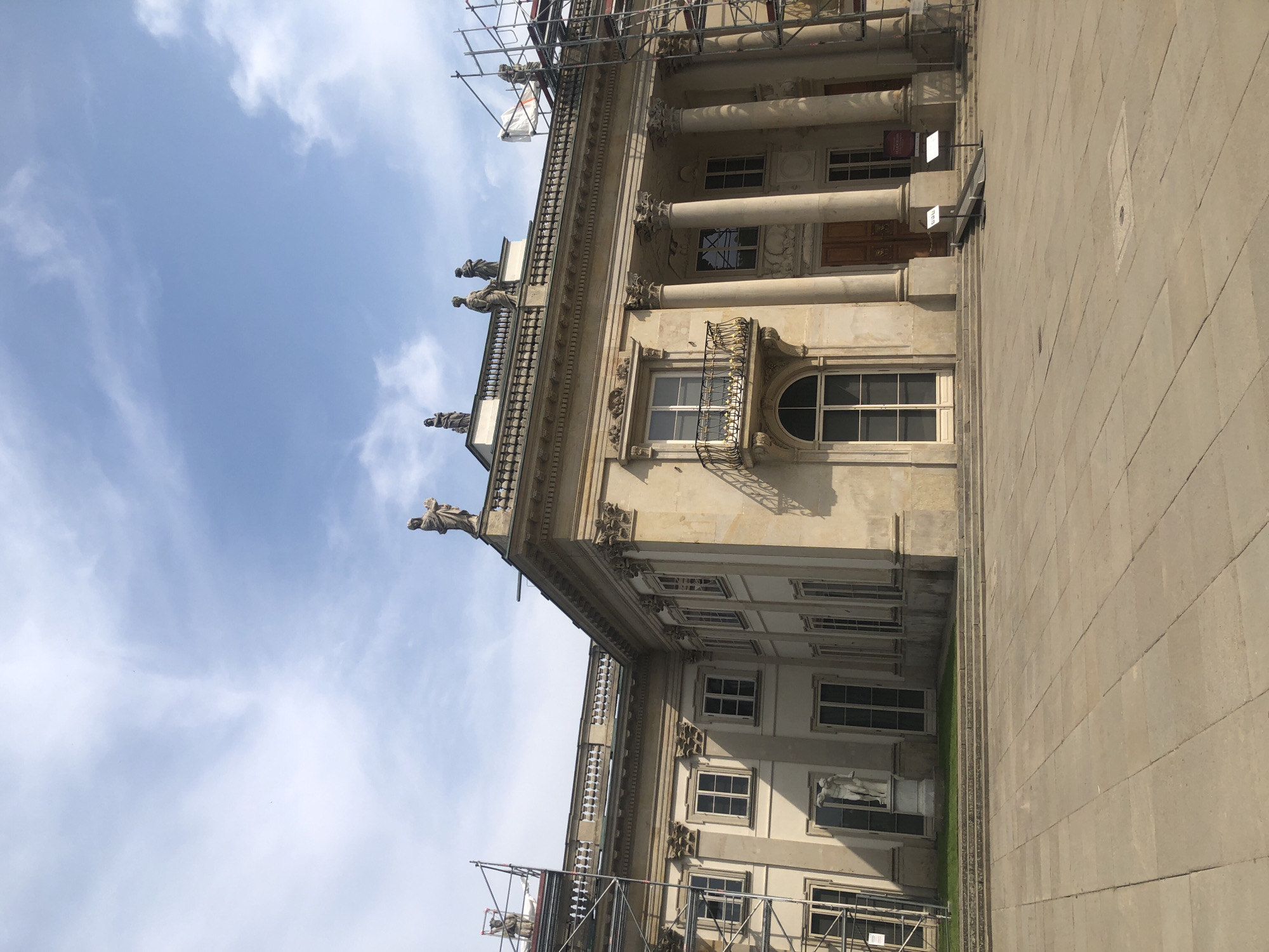 The image depicts the Royal Łazienki Palace in Warsaw, Poland. It is a daytime, outdoor shot with a bright, partly cloudy sky. The palace, constructed of light-colored stone, features classical architectural elements such as columns, a portico, and decorative sculptures on its roofline. Scaffolding is visible on the right side of the building, suggesting ongoing renovations or maintenance. In front of the palace is a large, paved courtyard with a set of stone steps leading up to the entrance. A statue of a male figure stands on a pedestal to the left, near the steps. Two information signs and a ramp are positioned in front of the main entrance. The overall scene is one of historical grandeur and architectural significance, with elements of preservation work in progress.