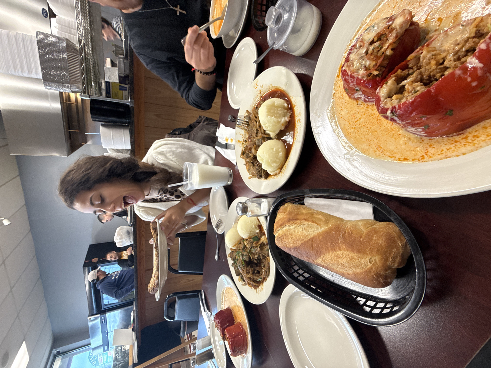 A woman is seated at a table in a restaurant, appearing to enjoy a meal. Various plates of food are arranged on the table, including stuffed peppers in sauce, meat with mashed potatoes, and a bread basket containing a loaf of bread. Other people are visible in the background, indicative of a dining establishment setting. The location is in Des Plaines, United States of America.