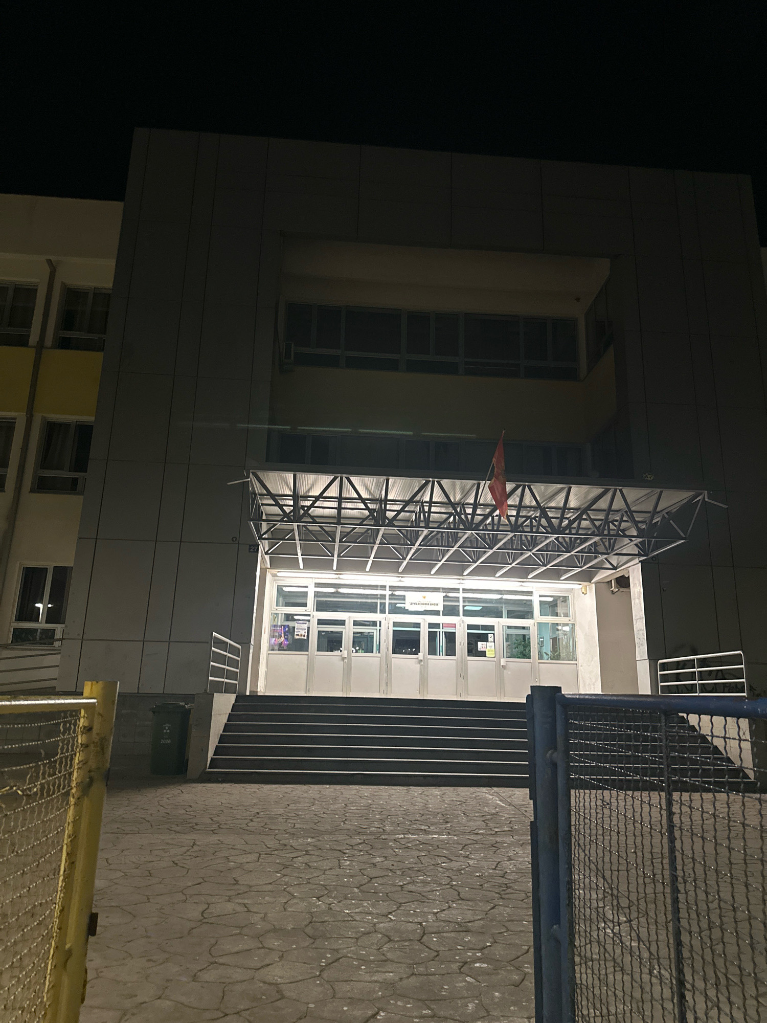 The image depicts the front entrance of a school building at night, likely located in Budva, Montenegro. The building features a modern, utilitarian architectural style with large windows and gray facade panels. A covered entryway with a metal frame structure extends over a set of wide stairs leading to double glass doors. A red flag is visible hanging from the roof of the entryway. To the left of the stairs, a green trash bin with the number "2026" is positioned on the paved walkway. The ground is covered in a patterned stone or concrete material. A metal fence is partially visible in the foreground, with a yellow post on the left and a dark metal fence on the right. The sky is dark, indicating it is nighttime, and the building is illuminated by artificial light.