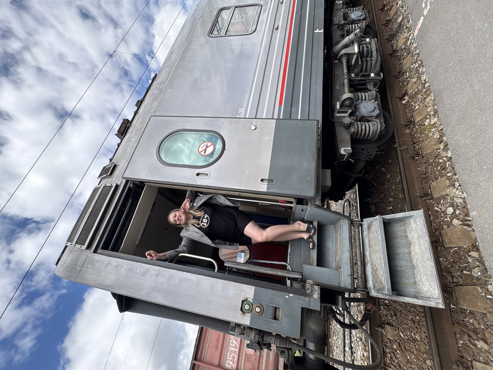A woman poses in the doorway of a train car in Rostov-on-Don, Russia. The train car is gray with red and white stripes. A "no smoking" symbol is visible on the door. The woman is wearing a black shirt with "18+" printed on it, black shorts, and a gray jacket. She is lying partly in and partly out of the doorway. The train is parked on railroad tracks next to a paved area. The sky, with white clouds, is visible behind the train.