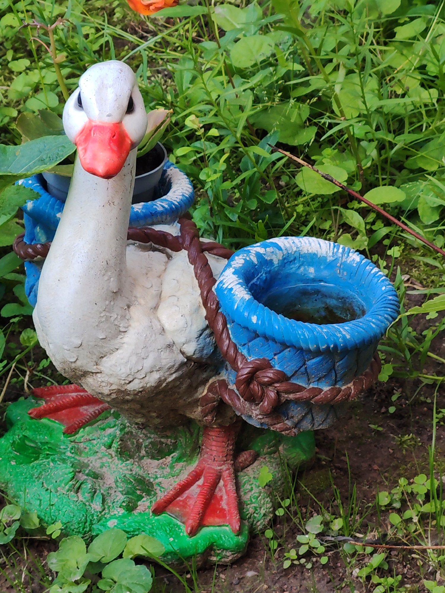 A white concrete statue of a duck, with red webbed feet and beak, stands in lush green foliage. The duck has two blue baskets attached to its back, held in place by brown rope. The statue is on a green, textured base. The background consists of dirt and various green plants and leaves. The scene is outdoors and appears to be in a garden or natural setting.