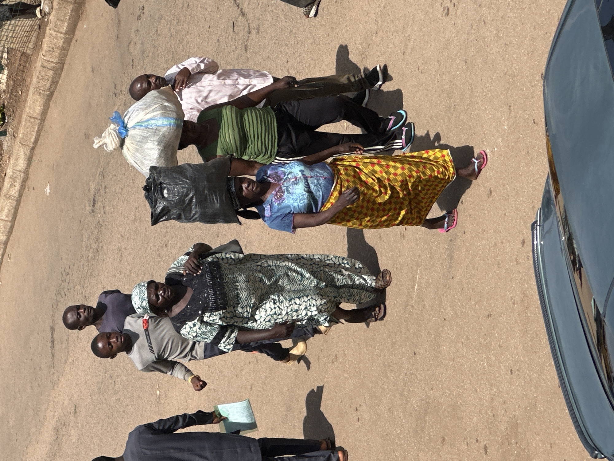 In Bukuru, Nigeria, a group of people are walking on a paved road. In the foreground, a person is carrying a large black bag on their head and a woman in a green and striped shirt is walking beside them. Behind them, a woman in a patterned dress is walking with a white bag on her head, followed by a boy in a gray shirt and a man in a dark shirt. Further back, another man in a striped shirt and dark pants is walking with the group. The environment is an outdoor urban setting with a slightly textured paved surface.
