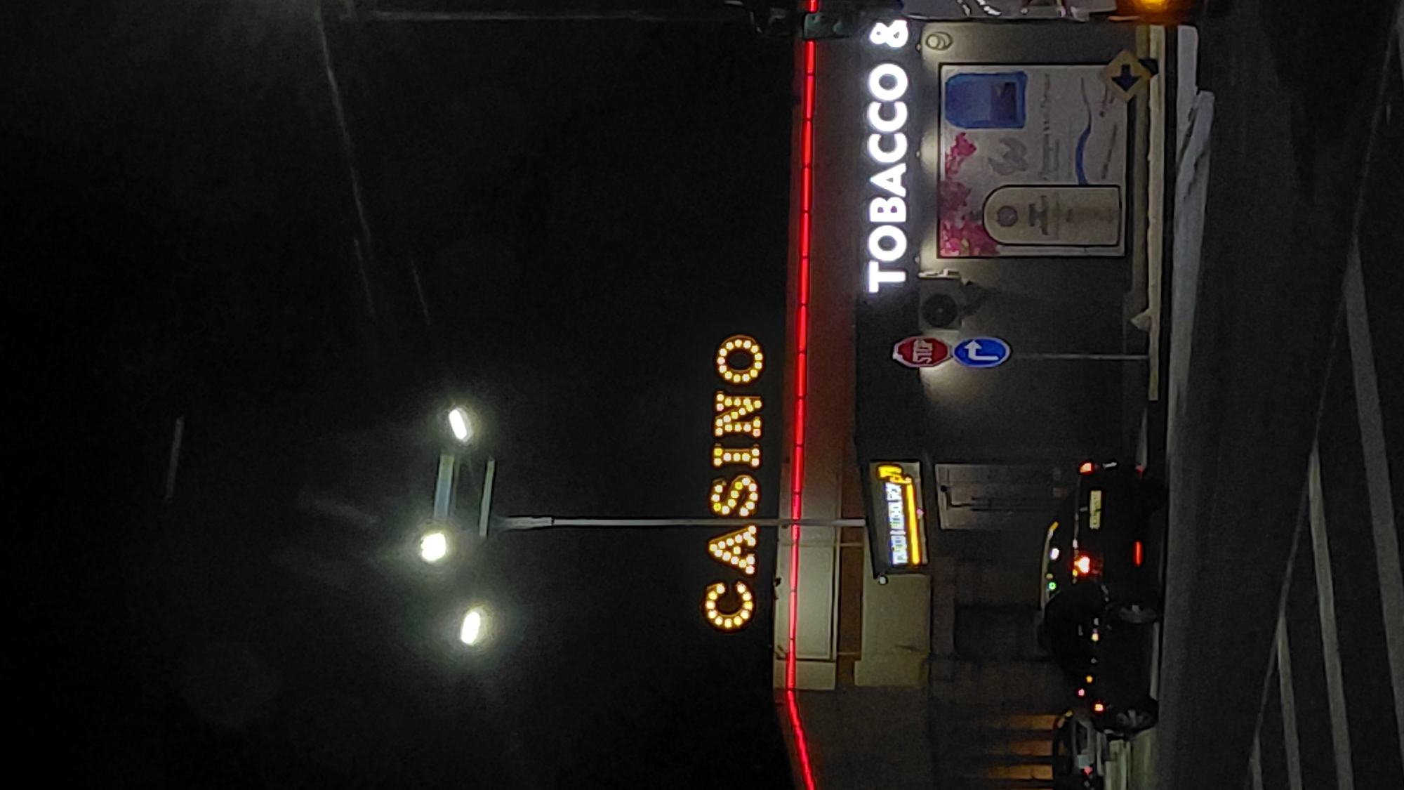 At night, a view of a casino and tobacco store in Unknown, Unknown, is depicted. Illuminated signs for "CASINO" and "TOBACCO &" are visible. A stop sign and traffic flow sign are also present. Several vehicles are on the road. A streetlight illuminates part of the scene.