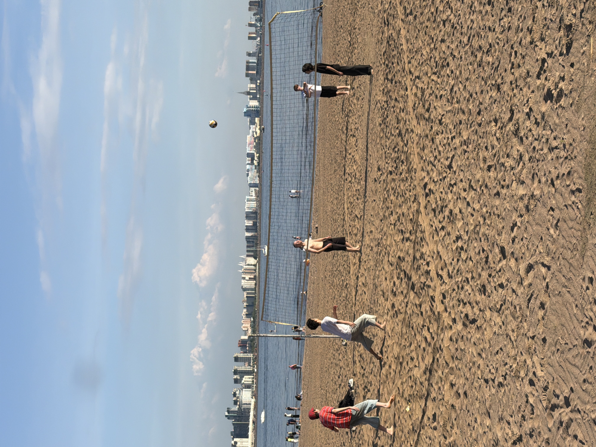 This media file captures a vibrant daytime scene on a sandy beach in Saint Petersburg, Russia, centered around a beach volleyball game.
The image shows a wide stretch of light brown sand, heavily textured with numerous footprints, indicating active use. A beach volleyball net stands across the frame, dividing the sandy court. Several individuals are engaged in the game or observing. In the foreground on the right, a person in a red plaid shirt and light shorts is seen bending low, seemingly in mid-play. Nearby, another person in a white t-shirt and light shorts walks towards the net. Further back on the right side of the net, a person with dark hair, wearing a dark top and dark pants, stands facing the court. On the opposite side of the net, a player in a white t-shirt and dark shorts is visible, along with another individual who is shirtless, suggesting sunny weather. A volleyball is suspended in the air above the net, indicating an active rally.
Beyond the sandy court, a calm body of water extends towards the horizon, likely the Gulf of Finland. A prominent city skyline stretches along the distant shore, featuring numerous buildings that suggest a bustling urban environment. The sky above is clear and bright blue, dotted with scattered white clouds, signifying pleasant and sunny weather conditions, likely during the late morning or afternoon. Several small boats or vessels are visible on the water in the distance. The overall scene conveys a leisurely and active atmosphere, with people enjoying sports and the outdoors. No visible text is present in the image.